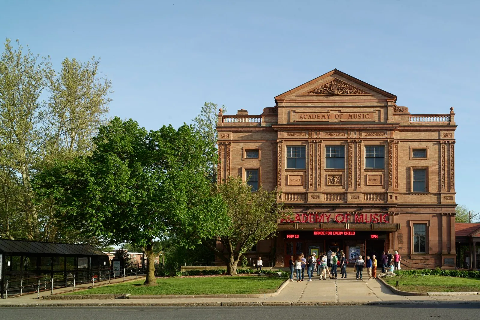 The Academy of Music Theatre on Main Street in Northampton on a sunny spring afternoon — a three-story Renaissance-revival facade in tan brick with sandstone trim, a pedimented central bay carrying the carved 'ACADEMY OF MUSIC' wordmark above a row of tall arched second-story windows, a red-lettered illuminated marquee over the ground-floor entrance with a small group of people gathered at the doors, a large leafy tree and a low bus shelter at the left edge, and a clear blue sky overhead.