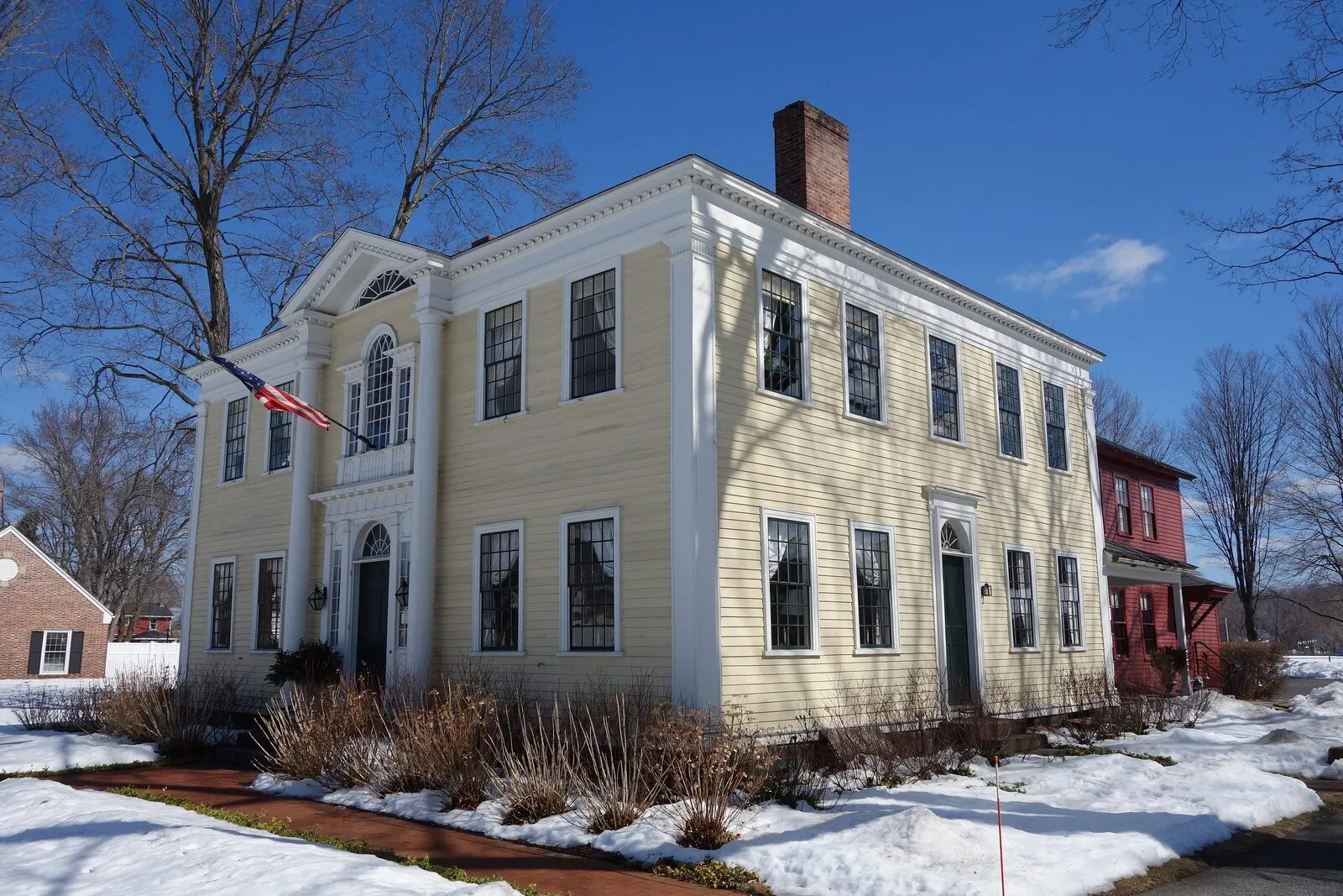 The Captain Charles Leonard House on Main Street in Agawam Center on a sunny late-winter afternoon: a two-story pale-yellow clapboard Federal house with white trim, a hipped roof, a deep entry portico flanked by pairs of slim Doric columns supporting a small entablature with a Palladian window above the door, an elliptical fanlight visible above the second-floor window, a US flag on a pole at the front, an adjacent red-brick service wing on the right, snow patches on the lawn and bare deciduous tree branches reaching up against the bright blue sky.