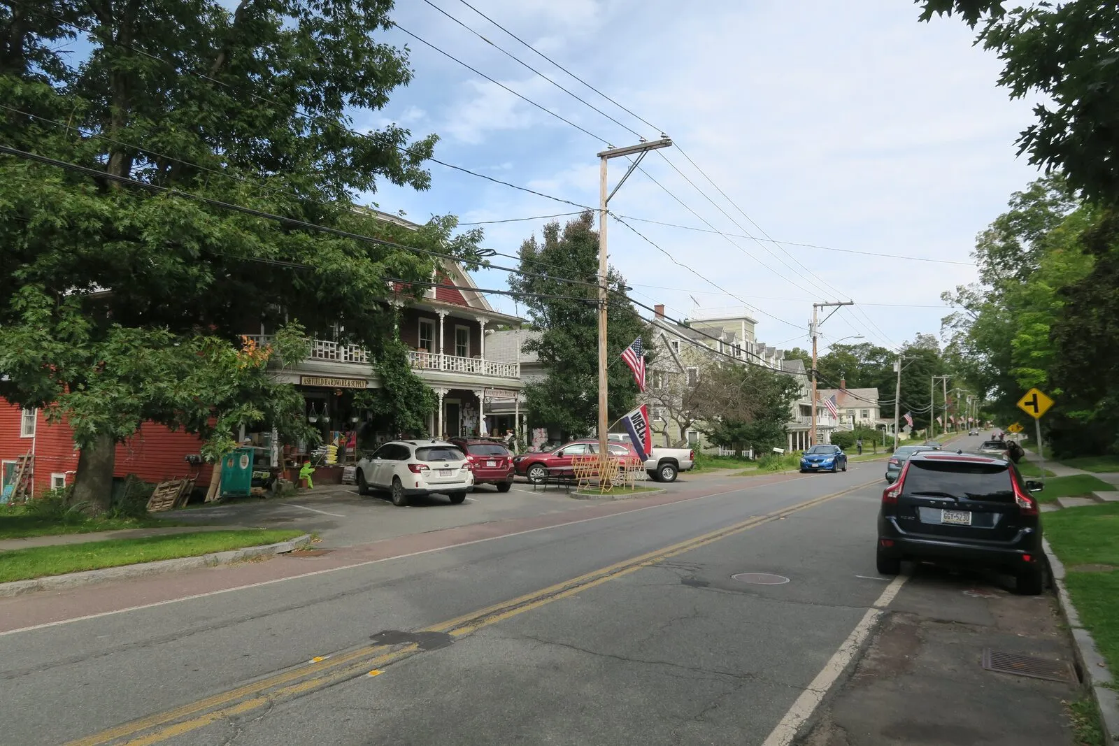 Looking west along Main Street (Route 116) through Ashfield Plain on a hazy summer afternoon: a long two-story clapboard porch-fronted commercial block on the left with an American flag hanging from the upper veranda, a small red shed and clapboard side-building set back at the very left, several cars angle-parked at the curb in front, a wooden telephone pole rising in the center foreground with overhead wires running across the frame, a yellow road-sign on the right shoulder, low clapboard houses and dark mature trees lining the right side of the road, the empty asphalt of Route 116 running into the distance under a pale blue sky with thin high clouds, and dense green foliage filling both edges of the view.