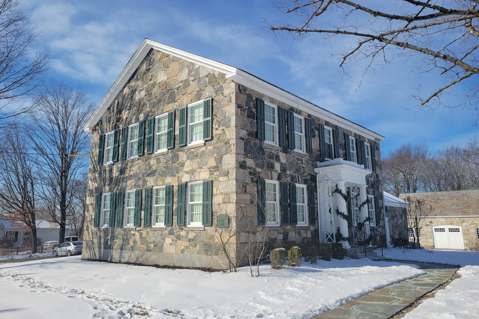 The Stone House Museum on the Belchertown common on a sunny January morning: a two-story 1827 fieldstone house with a steep gabled roof, mosaic of light tan and dark blue-gray stones in the walls, deep green shutters flanking the front-facing windows, a small classical entry portico over the front door, snow on the lawn and along the slate walkway leading to the door, bare deciduous tree branches reaching across the sky on the right, and a clear blue sky with scattered cumulus clouds overhead.