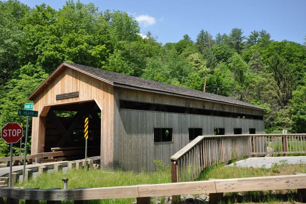 The Bissell Covered Bridge in Charlemont seen from the south on a sunny early-summer day: a 92-foot Long-truss covered bridge with vertical-board pine siding, a low-pitched dark asphalt-shingle gable roof, a pedimented portal facing the camera at left and the long flank of the bridge running away to the right with a row of small rectangular window openings, a stop sign and Park Street / N. Heath road signs at the near corner, a pedestrian walkway with a wooden rail along the near side, tall grass and Mill Brook's ravine in the foreground, and a wooded green hillside rising behind under a clear blue sky.