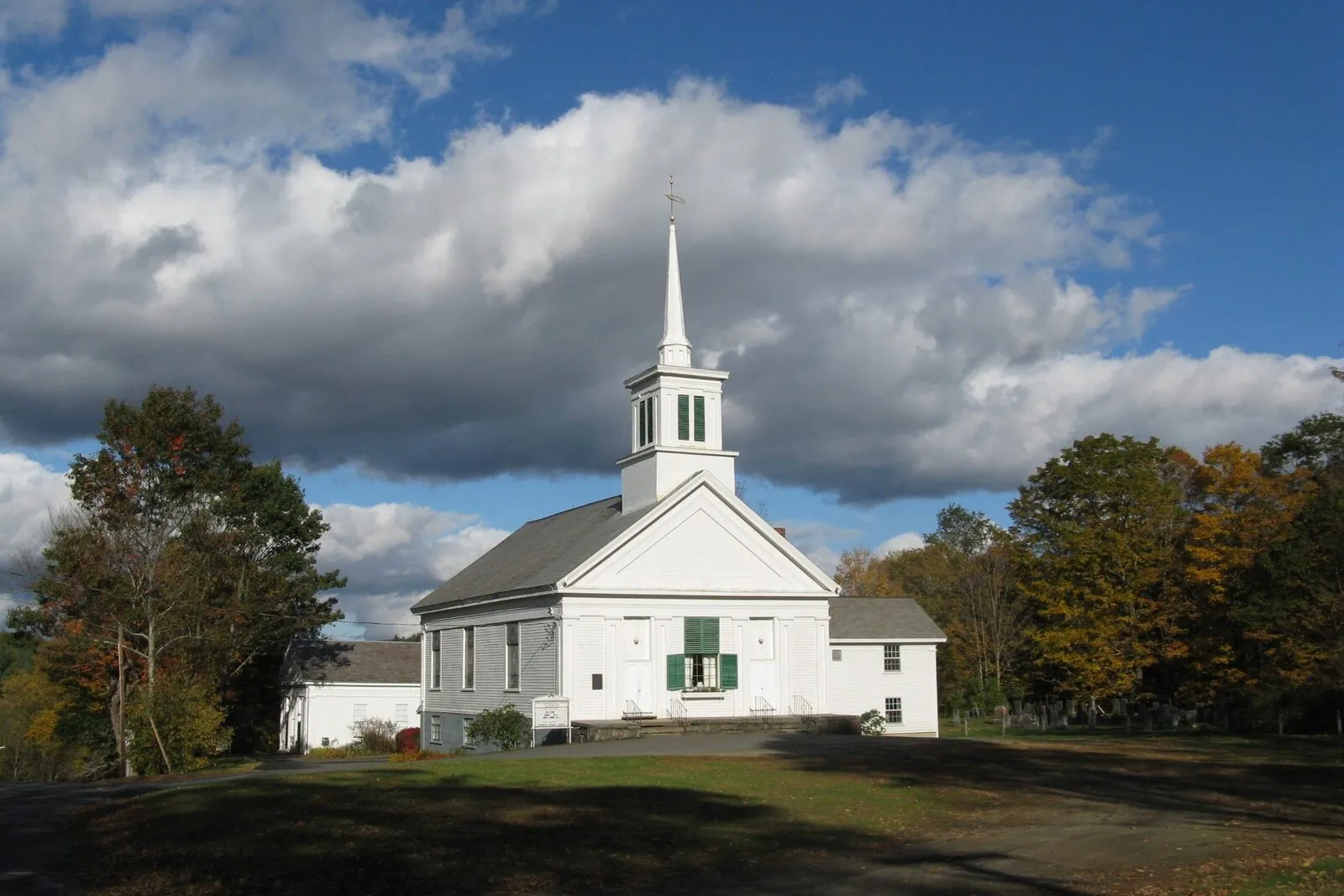 The First Congregational Church of Buckland on its hilltop in Buckland Center on a sunny October afternoon: a tall white-clapboard New England meetinghouse with a single classical pediment, a slim three-tiered spire rising to a gilded weather vane, two flanking single-story wings stepping down from the main block, deep-green-shuttered windows on the front, an old hipped-roof outbuilding tucked behind on the left, mature deciduous trees in mixed yellow-and-orange fall color framing the building on both sides, a sweeping green lawn in the foreground, and a dramatic deep-blue sky filled with bright white cumulus clouds overhead.