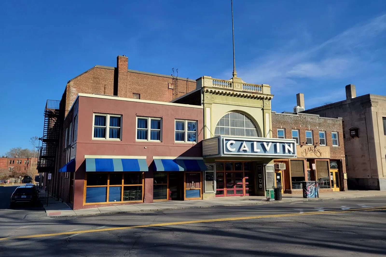 The Calvin Theatre at 19 King Street in downtown Northampton on a sunny winter afternoon — a two-story tan-and-brick commercial building on a corner, an ornate decorative pediment over the entry with 'CALVIN' lettered in white capitals on a blue marquee band, a tall vertical 'CALVIN' blade sign projecting from the building above the marquee, a row of blue-and-white striped retractable awnings over the street-level shopfronts to the left, side adjacent buildings flanking, and a few parked cars along the empty street under a clear blue sky with thin cirrus clouds.