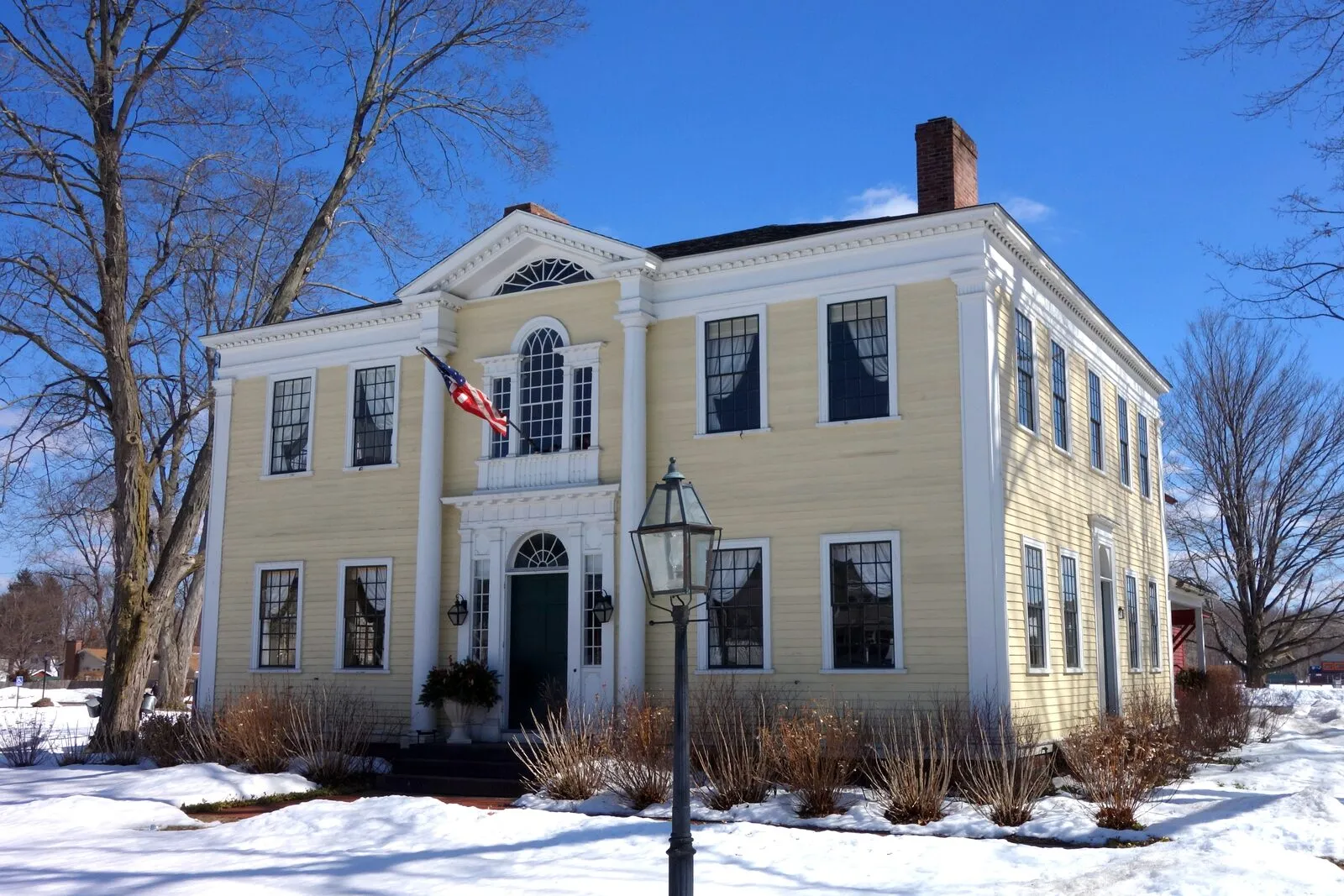 The Captain Charles Leonard House on Main Street in Agawam Center photographed from the front-left on a clear winter day. A two-story pale-yellow clapboard Federal mansion with white trim, a hipped roof, two pairs of two-story slim Doric columns flanking a recessed center entry with a green panelled door, a tall arched Palladian window centered above the entry under a fluted entablature, an elliptical fanlight visible at the gable end, white twelve-over-twelve sash windows on both stories, a brick chimney rising at the roof ridge, a black cast-iron lamppost in front, a U.S. flag on a wall-mounted pole beside the door, bare deciduous trees framing the lawn at left and right, snow on the ground, and a deep blue sky overhead.