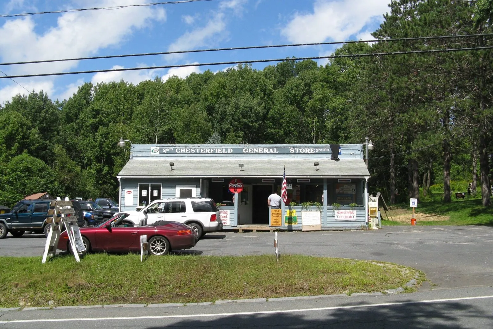 The Chesterfield General Store on a sunny late-summer afternoon — a long single-story clapboard building painted pale blue with white trim, a shallow gable-fronted porch over the central entry, a long horizontal black-and-white sign reading 'Chesterfield General Store' across the porch fascia, a Coca-Cola sign and an American flag flanking the entry, hand-lettered specials boards leaning against the porch posts, two parked cars in the gravel lot in front (a dark sedan on the left and a maroon convertible in the center), a wooden sandwich board at left, the empty two-lane town road sweeping in from lower right, mowed grass along the foundation, and a backdrop of mature deciduous trees in full green leaf under a deep-blue sky scattered with broken cumulus clouds.