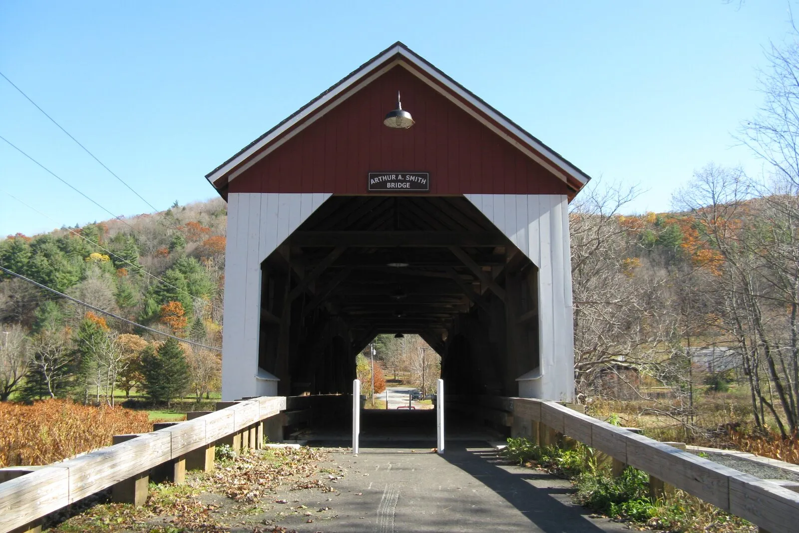 Looking through the east portal of the Arthur A. Smith Covered Bridge in Colrain on a clear October afternoon: a single-lane wooden covered bridge with red gable-end siding, white-painted side walls, and a small blue rectangular sign reading 'Arthur A. Smith Bridge' mounted on the front gable, a single bare bulb hanging beneath the gable peak, the dark interior of the timber Burr-truss tunnel framing a small distant view of the road and trees beyond, simple wooden plank guardrails running along both sides of a paved approach in the foreground, fall-color hardwoods and dark conifers rising on the hillside behind, and a deep cloudless blue sky overhead.
