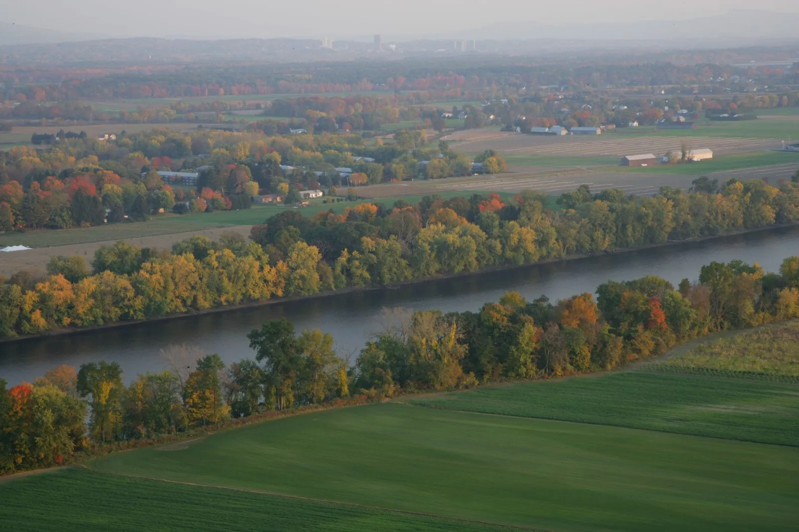 An aerial view of the Connecticut River winding through the Pioneer Valley on a hazy October afternoon — a long band of slow water curving across the middle of the frame between low forested banks tinted yellow, orange, and rust with peak fall color, broad green and tilled-tan farm fields stretching across the foreground and far bank, a few small white farm buildings scattered along a country road in the middle distance, and the low silhouette of distant hills along the horizon under a soft, even sky.