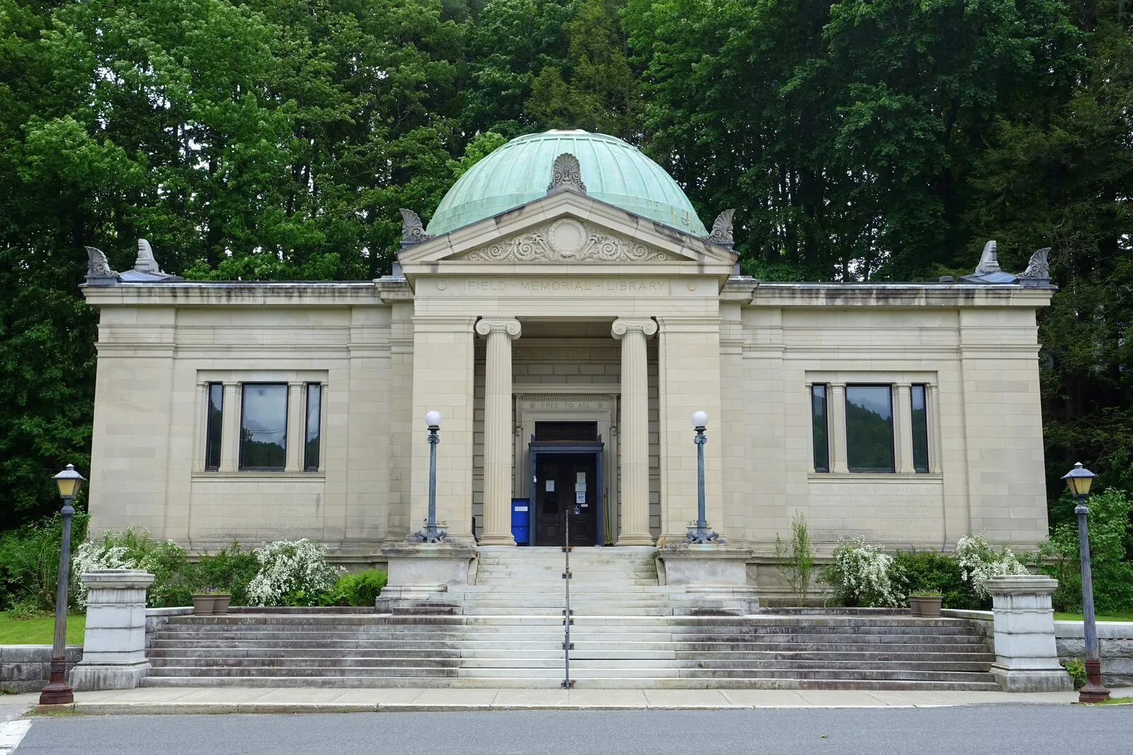 The Field Memorial Library on Main Street in Conway on a sunny early-summer afternoon: a Beaux-Arts limestone civic building with a low central pediment over a recessed entry flanked by paired Ionic columns, a tall verdigris-green copper dome rising over the central rotunda, low gable wings to either side with tall ground-floor sash windows, ornamental urns at the corners of the parapet, twin lampposts at the foot of the entry stairs, a black iron handrail running up the broad central staircase, neatly trimmed shrubbery flanking the foundation, and a backdrop of mature deciduous trees in full green leaf.