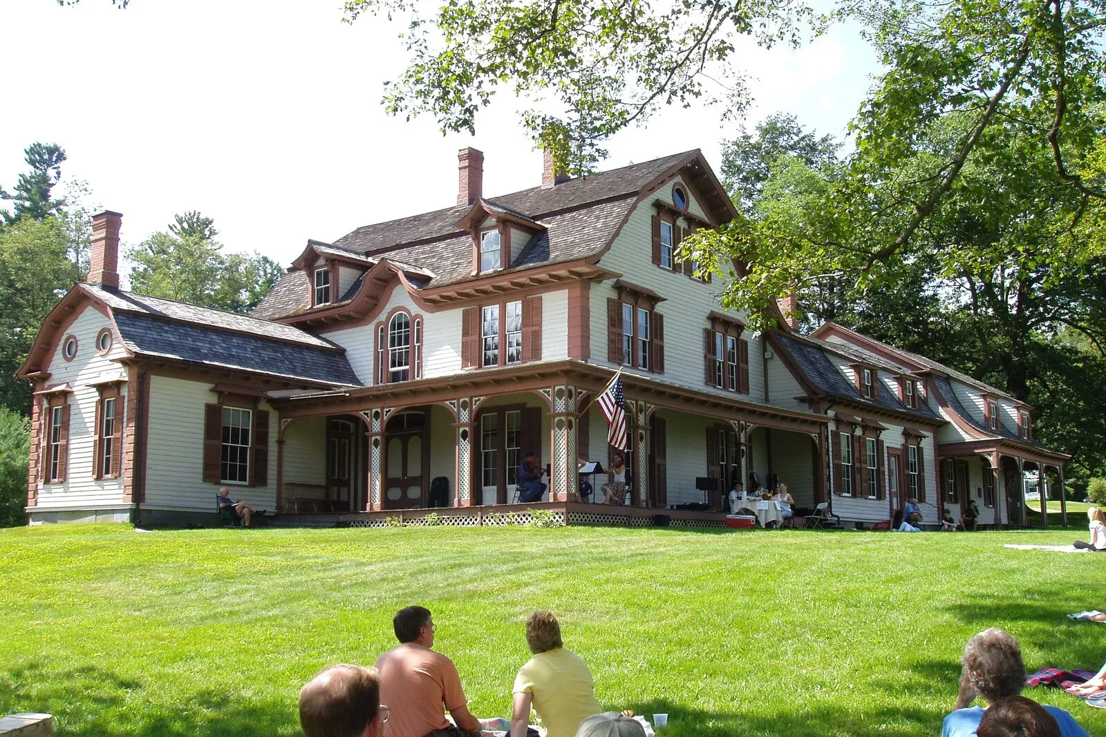 The William Cullen Bryant Homestead in Cummington on a sunny summer afternoon. A large two-and-a-half-story yellow-clapboard farmhouse with white trim and dark shutters, a steep gabled roof, two interior brick chimneys rising above the ridgeline, a long covered front porch running across the full width of the facade with slender white posts, a string quartet seated under the porch performing for visitors, a bay window projecting from the right side, low foundation plantings of shrubs and perennials along the front, a wide gravel driveway curving past the lawn, mature deciduous trees in full green leaf framing the house on both sides, and a clear blue sky overhead.