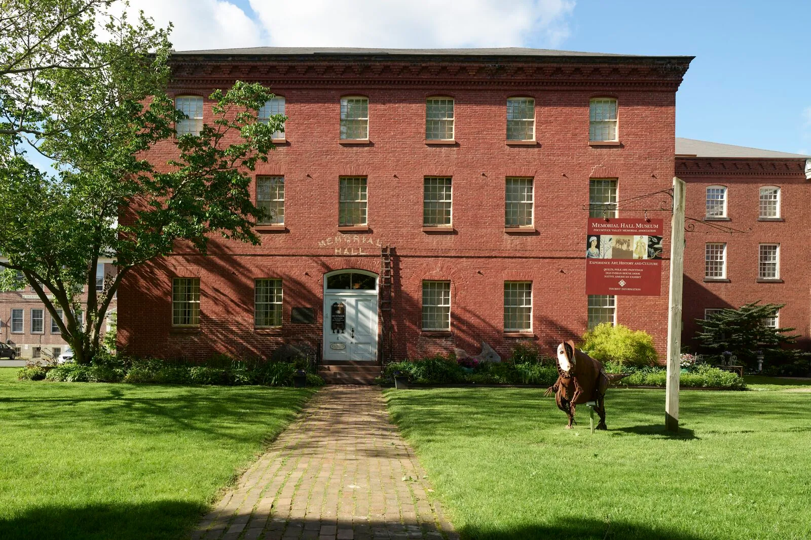 Memorial Hall in Old Deerfield on a sunny late-spring day — a three-story red-brick Federal building with white-trimmed windows and a hipped roof, 'MEMORIAL HALL' lettered across the facade above the entry, a brick walkway leading from the lawn to the white-framed door, a cast bronze figure on a pedestal at right, mature shade trees flanking the building, green grass in the foreground, and a clear blue sky with a few light clouds.