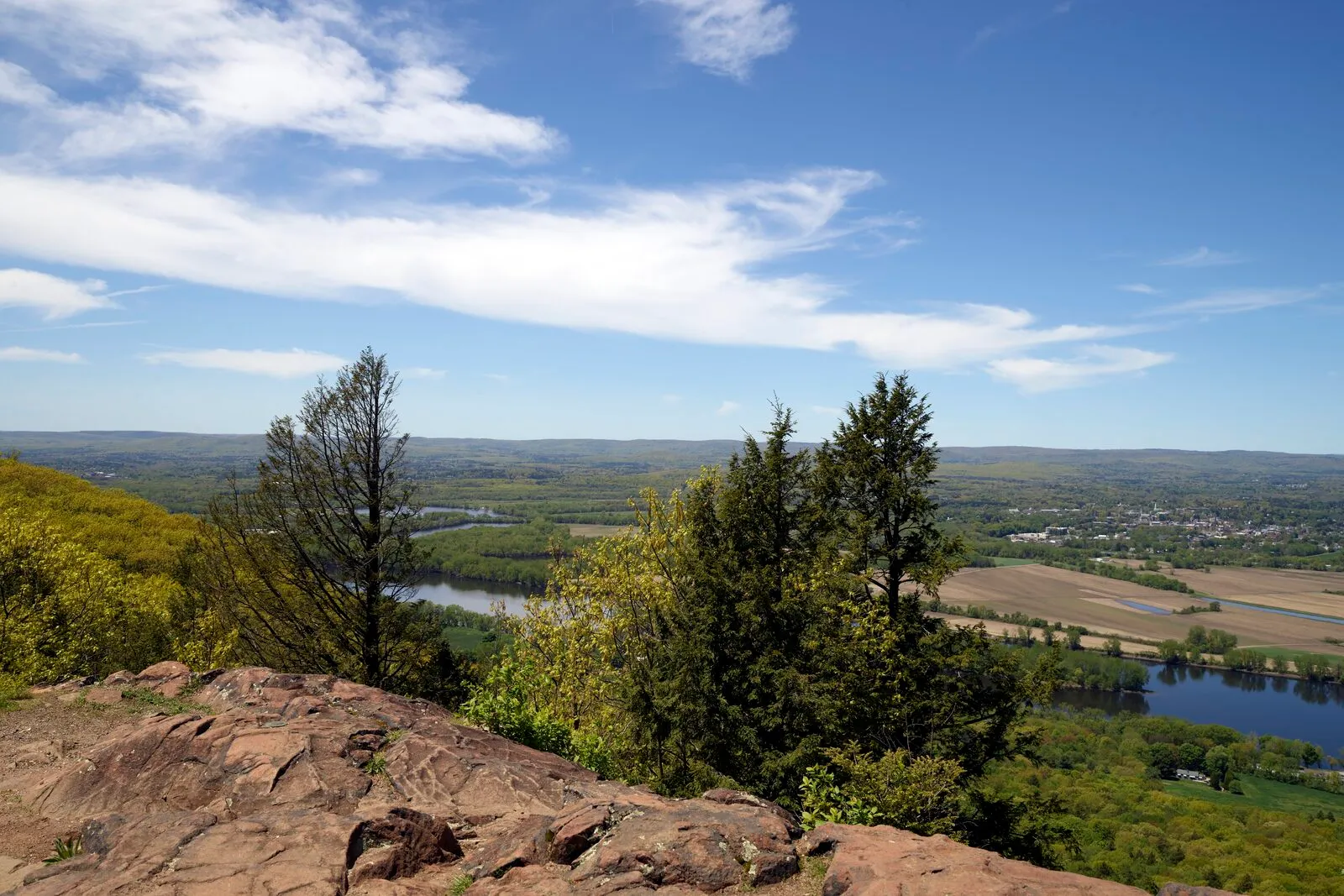 The Connecticut River Valley seen from the summit of Mount Holyoke in Skinner State Park on a clear May morning — exposed reddish-brown trap-rock bedrock in the foreground, a few young leafed-out deciduous trees mid-frame, the broad Connecticut River winding through patchwork farmland and small wooded blocks below, a low band of distant hills along the horizon, and a bright blue sky with thin streamers of cirrus cloud overhead.