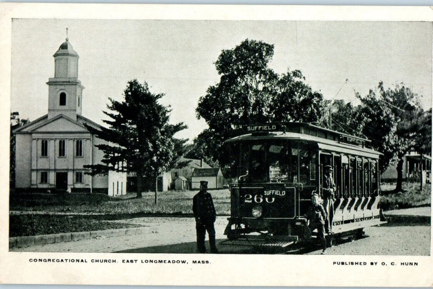 A black-and-white printed postcard from the early 1900s captioned 'CONGREGATIONAL CHURCH, EAST LONGMEADOW, MASS.' showing a wide unpaved village street with a tall white-clapboard Greek-Revival meetinghouse with a square bell-tower and lantern cupola at left, a tall deciduous tree in full leaf rising over a small white outbuilding behind it, and at right an open electric streetcar (destination sign 'SUFFIELD' and car number 266) stopped at a switch with two uniformed conductors in dark coats and peaked caps standing in the road, a row of trees and small wooden buildings receding behind it. Captioned in the lower right 'PUBLISHED BY O. C. HUNN.'