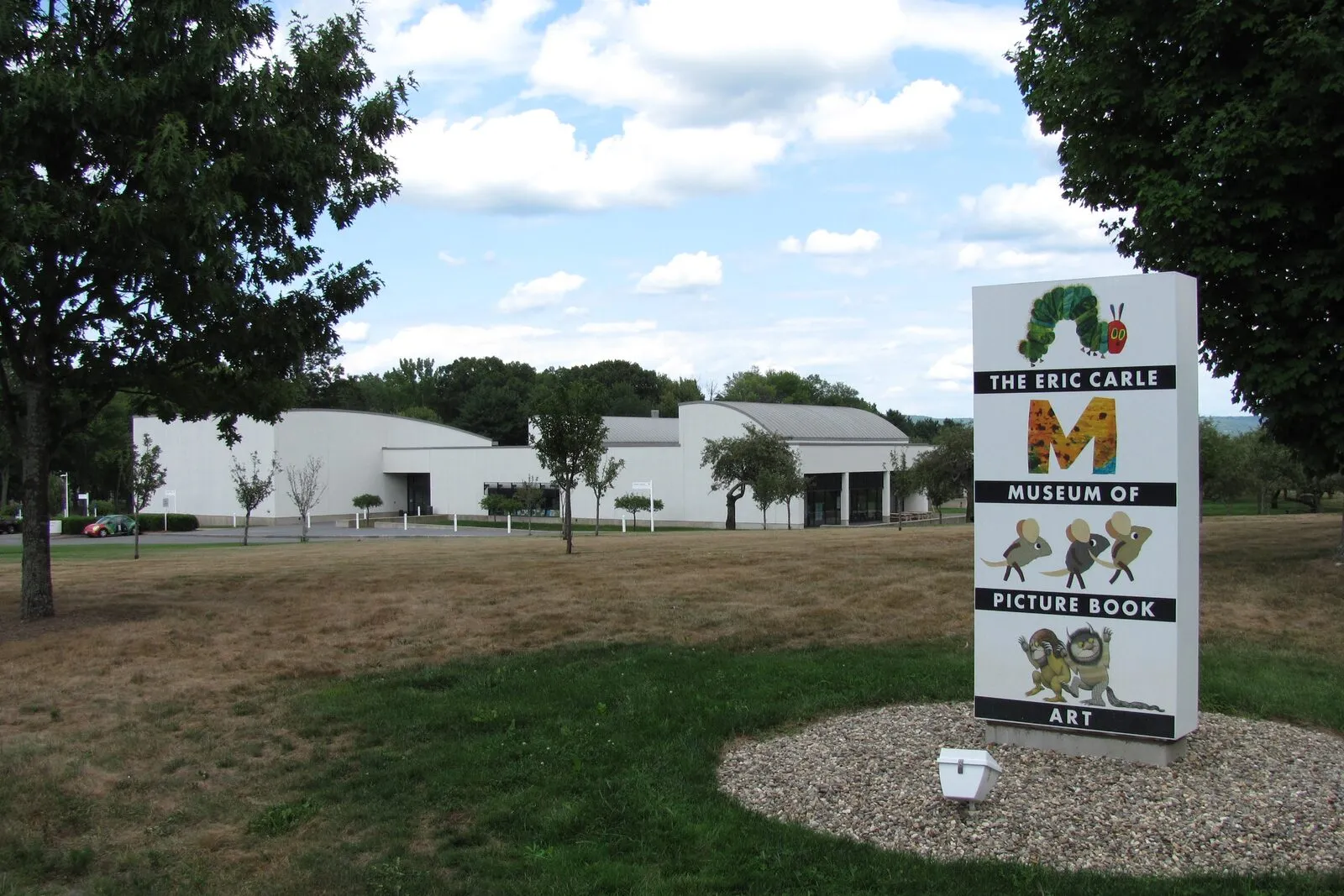 The Eric Carle Museum of Picture Book Art on a summer afternoon: a low, flat-roofed white building set on a wide open lawn with scattered young trees, and a tall white pillar-sign in the right foreground carrying the Very Hungry Caterpillar illustration above the stacked words THE ERIC CARLE / MUSEUM OF / PICTURE BOOK / ART, with bright white clouds in a blue sky behind.
