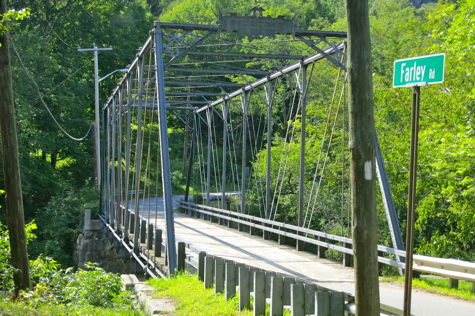 The Farley Road Bridge in the Farley village of Erving on a sunny summer afternoon: a one-lane 19th-century iron truss bridge with riveted dark-gray Phoenix-column posts, diagonal eyebars, and an X-pattern of overhead bracing, spanning the Millers River from a stone abutment in the foreground across to the far bank, a worn timber-plank deck running between low wooden curbs, dense green deciduous and conifer foliage filling both banks, a green Farley Rd street-name sign on a metal pole at right next to a wooden utility pole and a low galvanized guardrail, and patches of sunlight cutting across the truss.
