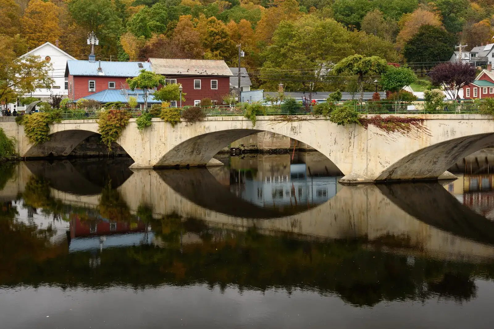 A stone arch bridge in Greenfield with peak fall foliage on the surrounding hills, reflected in the still water below.
