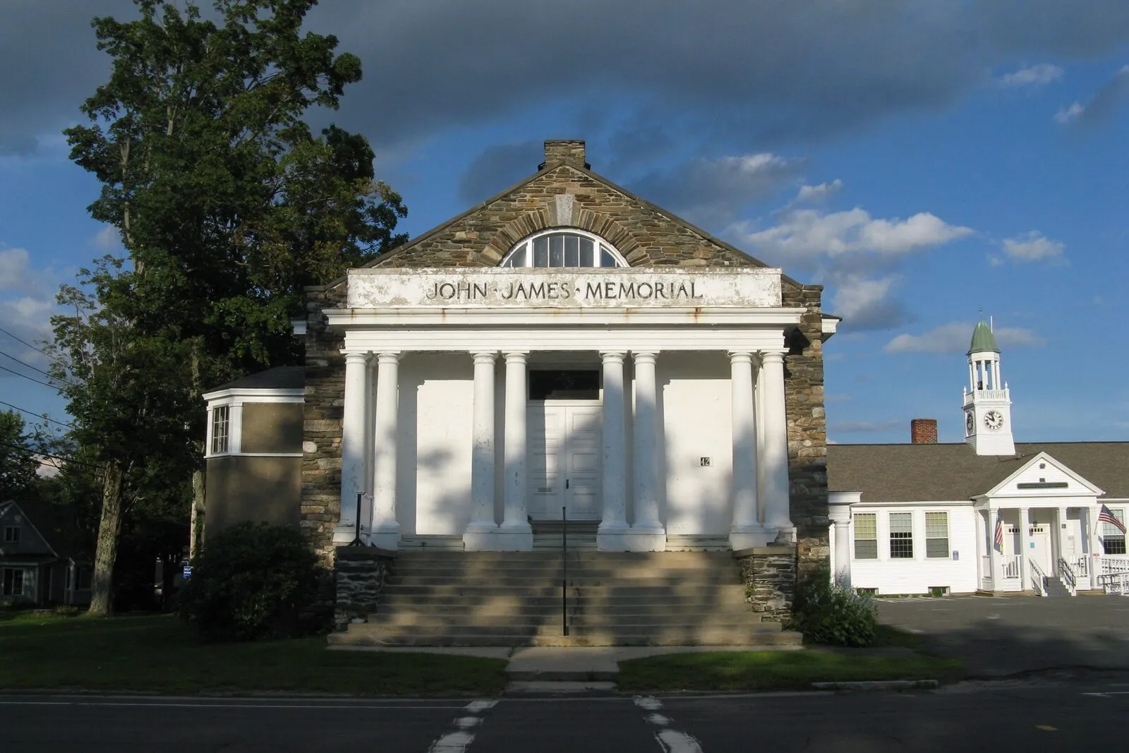 The John James Memorial building in Goshen Center on a clear summer afternoon. A two-story civic hall in locally-quarried rough fieldstone with deep-set window openings, a steeply-pitched gable roof in dark slate-gray asphalt shingles, a small projecting entry porch with a low gabled hood over double wooden doors, paired ground-floor sash windows trimmed in white, a small bell-cote turret rising at the front-left corner of the roofline, a thin metal flagpole at the foundation line, mature deciduous trees in full green leaf framing the building on both sides, and a strip of mown lawn running down to the road in the foreground.
