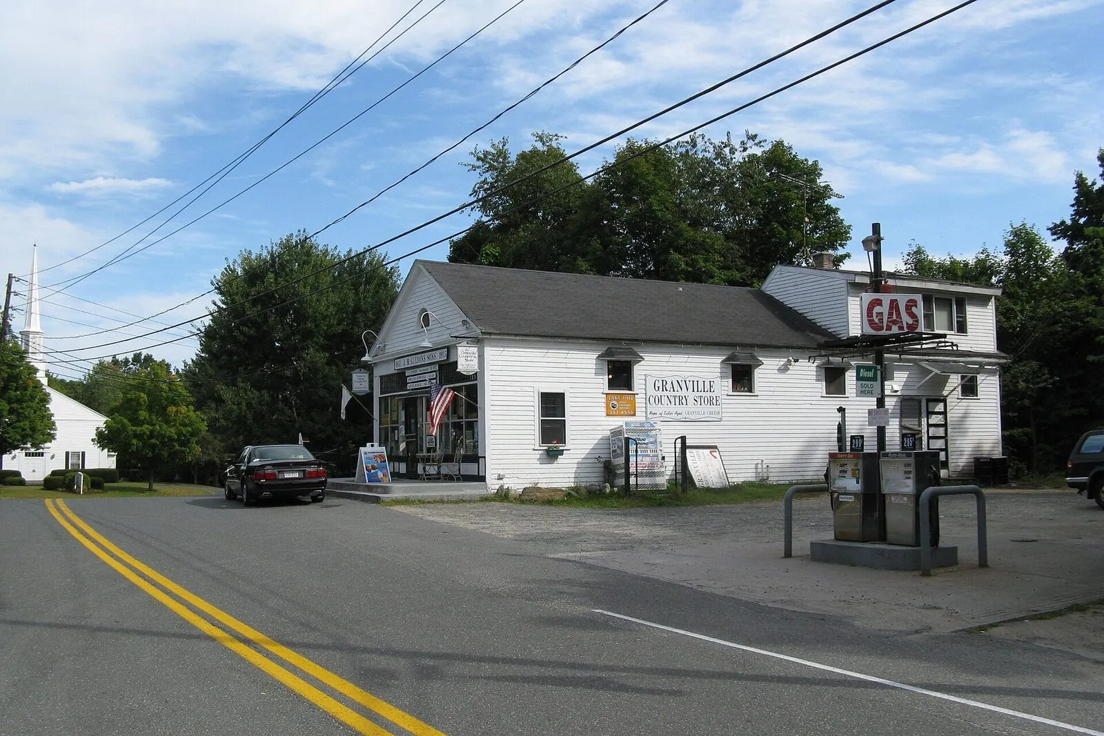 The Granville Country Store on a sunny late-summer afternoon, viewed from the road on the south side: a long white-clapboard two-story building with a gable-fronted main block on the left, a smaller attached single-story store wing in the middle, and a tall illuminated red-and-white GAS sign on a pole at right above two old-style gas pumps under a small canopy, an American flag hanging on the porch, hand-painted signage on the storefront reading 'Granville Country Store,' a single dark sedan parked in front, deciduous trees framing the lot, the double yellow line of the rural state highway curving past in the foreground, and a blue sky with scattered cumulus clouds overhead.