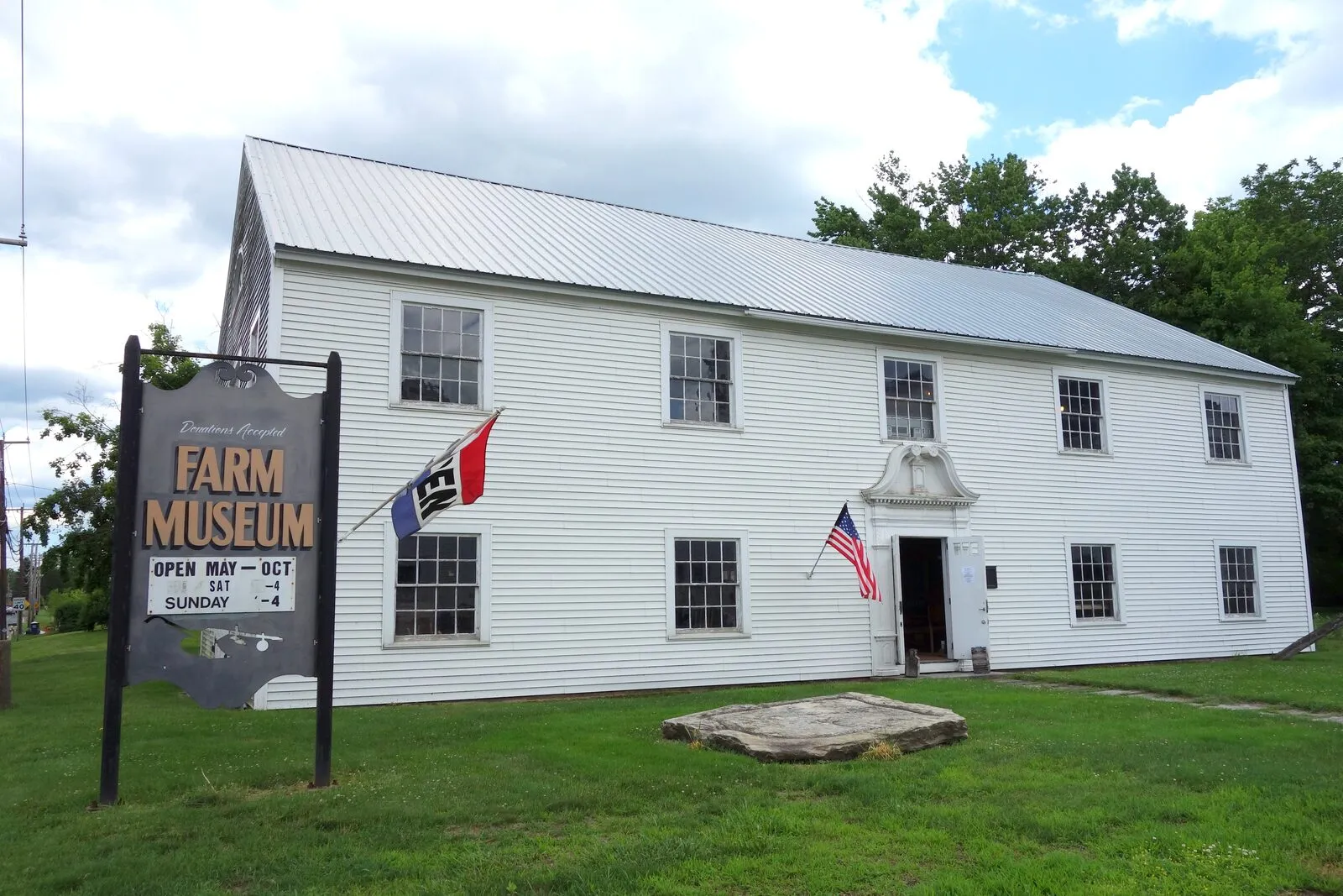 The Hadley Farm Museum, a long two-story white-clapboard barn with a standing-seam metal roof, photographed in summer from the lawn. A black sandwich-board sign at left reads 'FARM MUSEUM · OPEN MAY–OCT · SAT & SUNDAY 1–4', a tall flagpole flying the US flag stands center, an ornate colonial doorway with a small pediment is recessed under a hood, six-over-six sash windows run in pairs across the upper and lower stories, a New England 'Don't Tread on Me' style flag hangs to the left of the entrance, a granite mounting block sits on the lawn in front, and a leafy tree-line forms the backdrop under a partly cloudy sky.