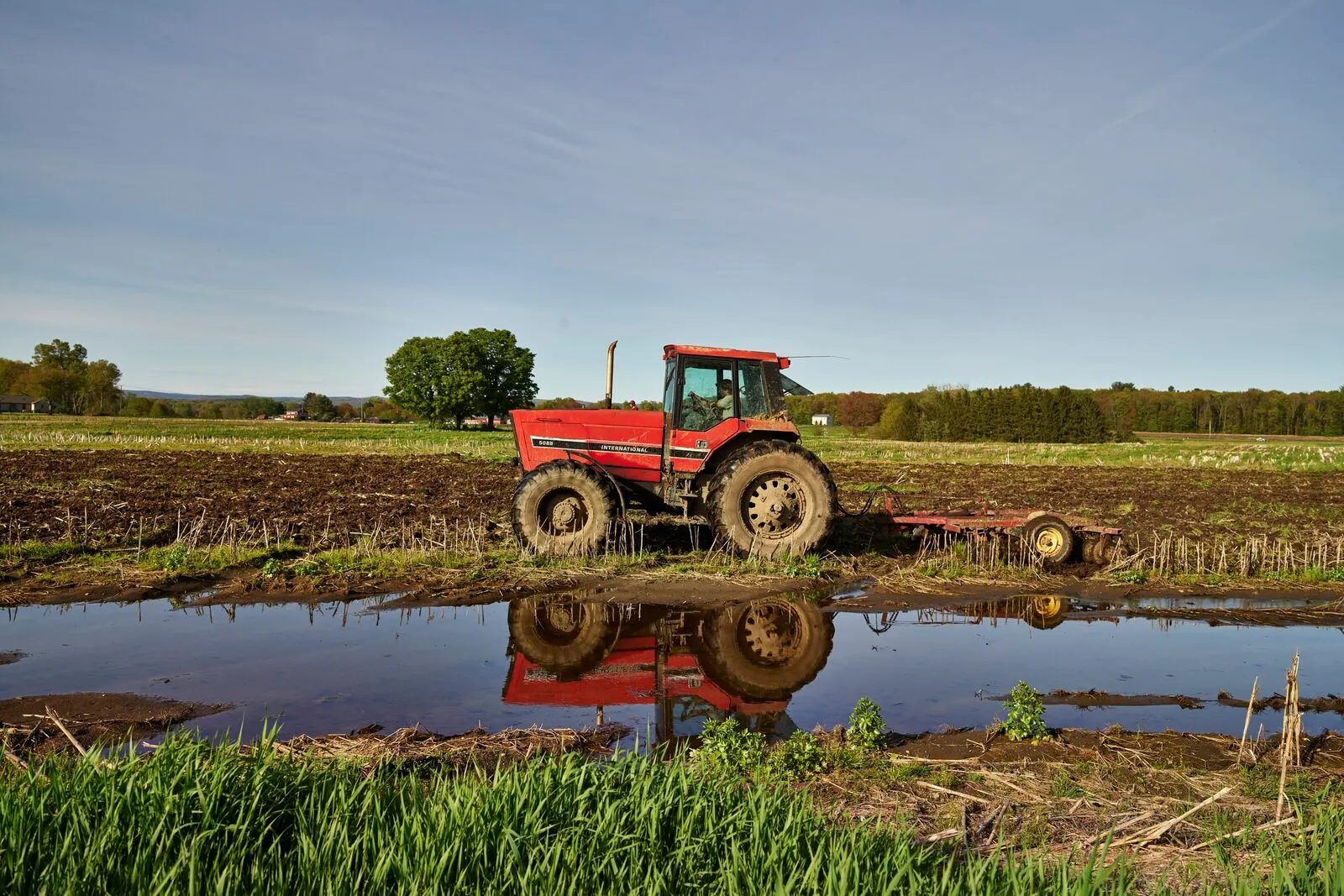 A red farm tractor and field-cultivator at the edge of a freshly tilled cornfield in Hadley on a clear evening in late spring: a long puddle of standing water in the foreground reflects the tractor and a band of grass, dark furrowed soil stretches across the middle of the frame, a hedgerow and distant tree line cross the background, and the low hills of Pelham are visible on the horizon under a clear blue sky.
