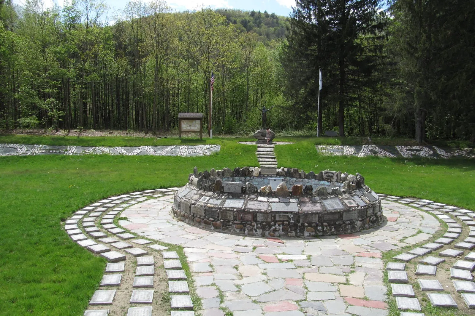 The Hail to the Sunrise memorial at Mohawk Park in Charlemont on a sunny spring afternoon. The bronze statue of a Native warrior with arms uplifted stands in the middle distance behind a low circular ring of about a hundred inscribed stones around a small reflecting pool, with a stone walkway in the foreground, a wooden interpretive sign and U.S. flag to the left, a tall Christmas-tree-shaped conifer to the right, and the wooded ridge of the Deerfield River valley rising under a partly cloudy sky behind.