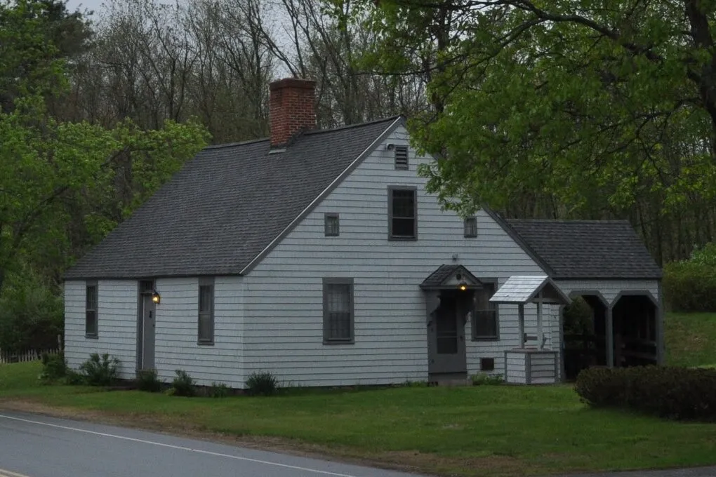 The Thornton W. Burgess House on Main Street in Hampden, photographed in 2014 — a long, low, two-story white-clapboard 19th-century farmhouse with a steeply pitched dark-shingled roof, several brick chimneys along the ridgeline, multiple gabled wall dormers, two-over-two and six-over-six sash windows, an attached white side ell stretching out to the right, a long covered front porch with simple square wooden posts running across most of the façade, and a clipped green lawn with a few small shrubs and a paved walkway in front of the house, framed by mature deciduous trees in full summer leaf with a hazy bright sky overhead.