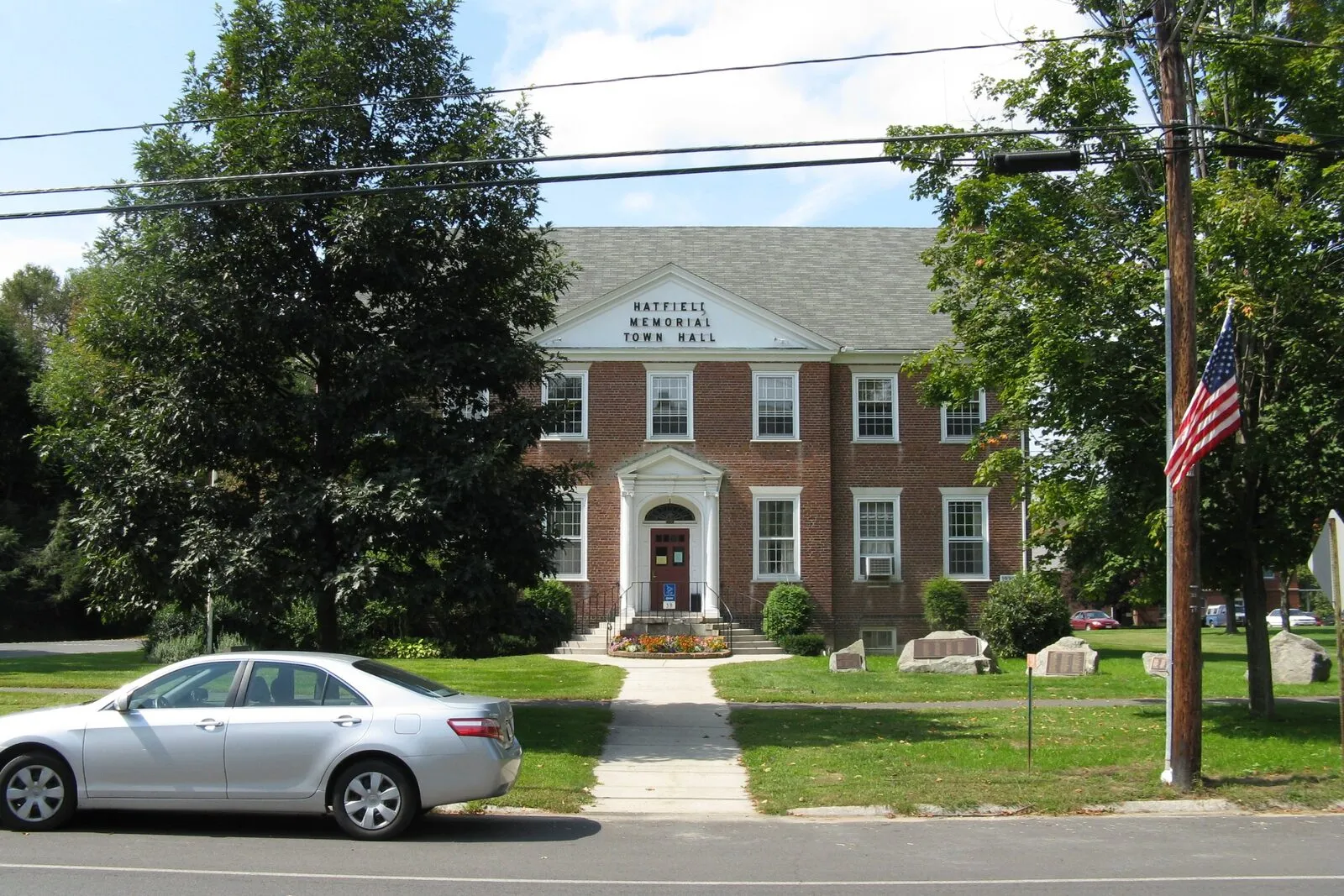 The Hatfield Memorial Town Hall on Main Street on a sunny late-summer afternoon: a three-bay red-brick civic building with a low pitched roof and broad cornice, six tall ground-floor sash windows flanking a recessed center entry with a small white-trimmed gable above lettered 'HATFIELD MEMORIAL TOWN HALL', stone foundation, a US flag flying from a pole at right, three large boulders set in the front lawn, mature street trees framing the building, and a single silver sedan parked at the curb in the foreground.
