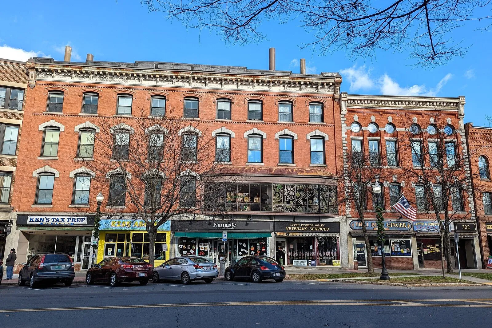 A late-fall view of the 278–302 block of Main Street in downtown Greenfield on a clear afternoon. A continuous row of three- and four-story 19th-century brick commercial buildings with painted cornices and ground-floor storefronts running along the sidewalk, the restored Hawks & Reed building among them with its tall round-arched second-story windows, mature street trees with bare branches lining the curb in the foreground, parked cars along the meter strip, and a clear blue sky above.