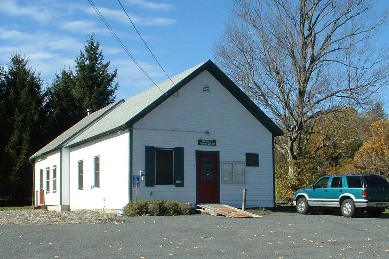 The Hawley Town Hall on a clear fall afternoon — a small one-and-a-half-story white-clapboard front-gabled building with a low pitched roof, a single red entrance door at center facing a wooden ramp, two pairs of shuttered double-hung windows flanking the door, a small framed bulletin board mounted on the right side of the front wall, dark conifers crowding behind on the left, a leafless deciduous tree to the right with a band of orange autumn foliage in the distance, a teal SUV parked at right on a wide gravel-and-asphalt lot scattered with dry leaves, overhead power lines crossing the deep-blue cloudless sky.