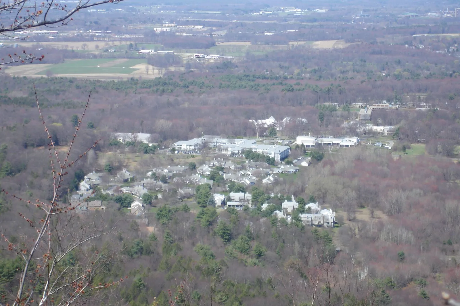 A view north from the summit of Bare Mountain in the Holyoke Range on a flat-light early-spring day — bare-branched deciduous forest in the immediate foreground, the white-roofed buildings of Hampshire College clustered in the middle distance, the broad open Pioneer Valley spreading north with farmland and a distant cluster of college and town buildings, and a low haze of forested ridges receding to the horizon under an overcast sky.