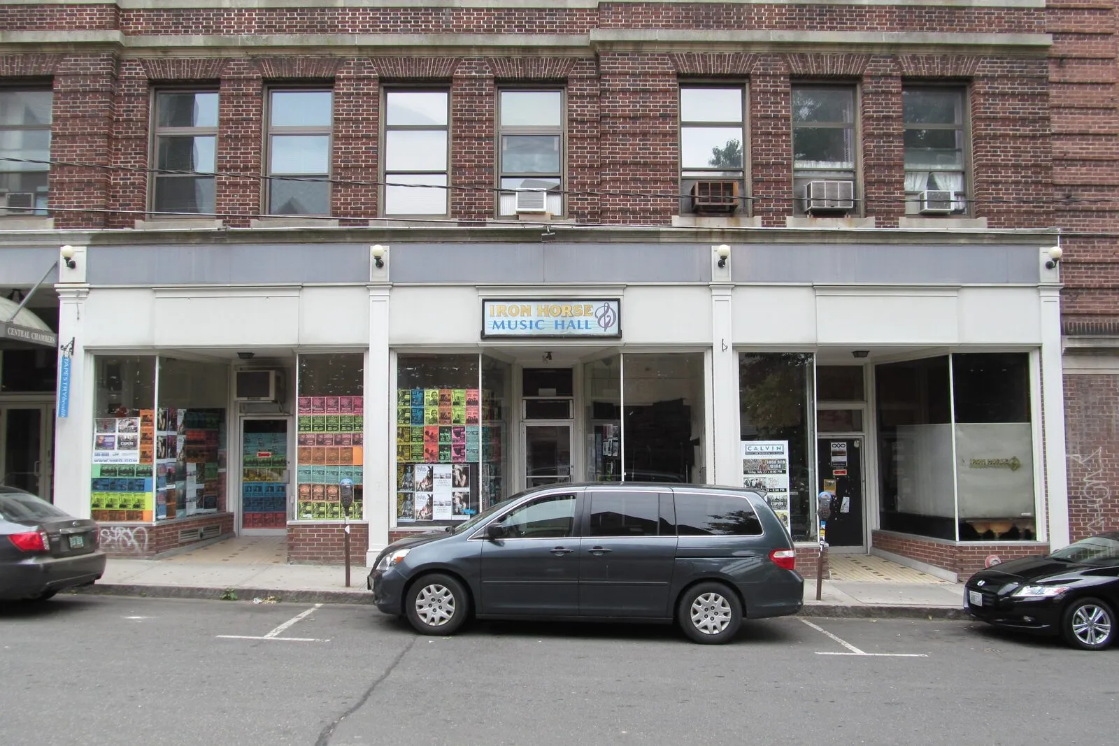 The Iron Horse Music Hall storefront on Center Street in Northampton on an overcast summer afternoon. A red-brick four-story commercial building with double-hung white-trimmed windows on the upper floors and several through-the-wall air conditioners, a continuous grey awning running across the ground floor, and a small painted sign over the central glass entry door reading 'Iron Horse Music Hall' beside a yellow musical-note logo, flanking display windows filled with concert posters and flyers, an empty white-painted glass storefront on the right, a grey minivan and a black sedan parked head-in at metered spaces along the curb, and a sliver of green street tree visible above the right end of the building.
