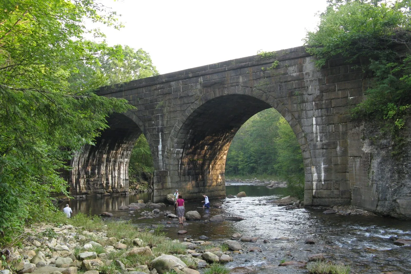 A double-span dry-stone railroad arch bridge over the West Branch of the Westfield River near Chester, Massachusetts. The bridge is built of large hand-cut granite blocks weathered grey, with two tall rounded arches spanning a shallow rocky river. A few small figures wade in the water beneath the arches; trees in full summer leaf rise on either side.