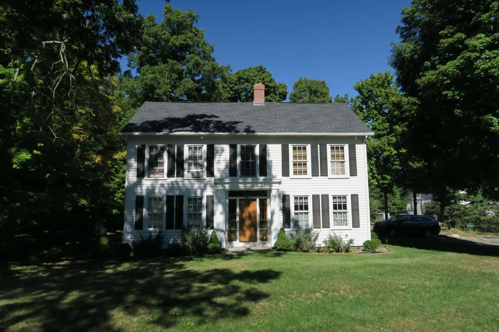 The Increase Sikes House on Center Street in Ludlow Center on a sunny late-September afternoon: a two-and-a-half-story white-clapboard center-chimney New England colonial with a side-gable roof, a single tall brick chimney rising through the ridge, dark green louvered shutters flanking the six-over-six sash windows on both stories, a recessed center entry with a panelled door under a simple pediment, a low foundation of granite block, a shallow front lawn in summer green, foundation plantings of small evergreen shrubs along the front, mature deciduous trees in full leaf framing the house at left and right and arching overhead, and a deep blue sky with thin scattered clouds visible above the canopy.