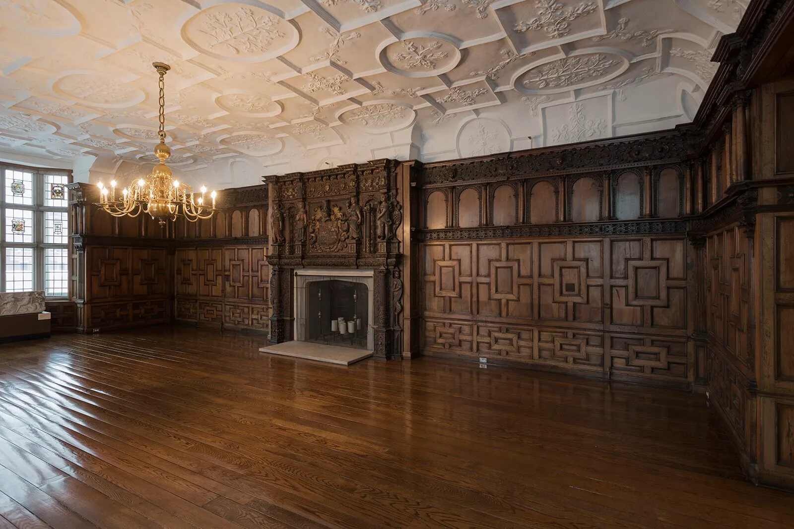 The Rotherwas Room at the Mead Art Museum, a 17th-century English Jacobean room reinstalled inside the museum, with dark oak paneling carved in geometric strapwork covering the walls, an ornate Tudor stone chimneypiece centered on the back wall, leaded-glass casement windows on the left wall, an intricate plaster ceiling of cartouche medallions overhead, a brass chandelier hanging mid-room, and a polished dark wood floor.