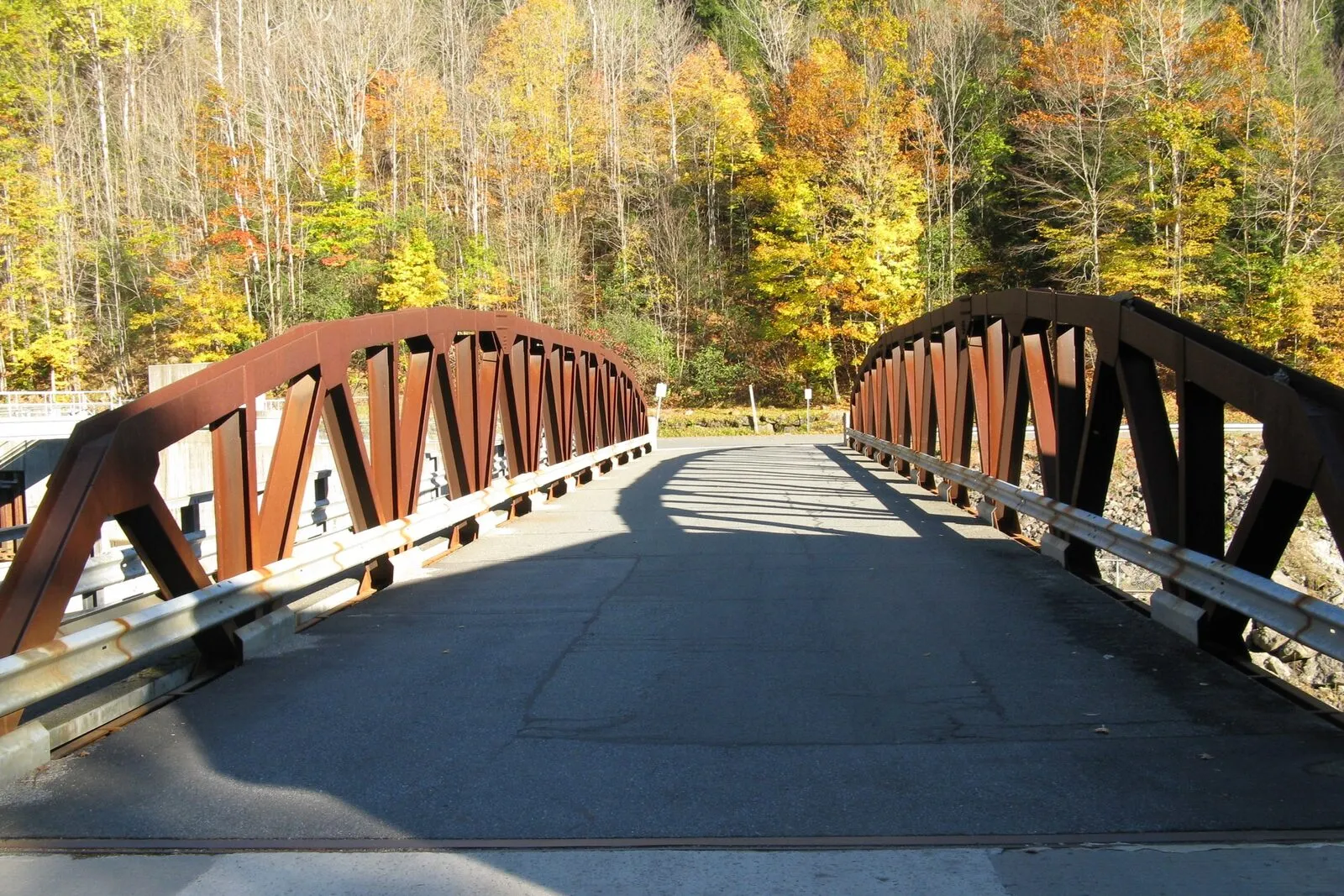 Looking south across a small steel pony-truss highway bridge spanning the Deerfield River at the village of Monroe Bridge on a sunny fall afternoon. The rust-orange painted bridge has open lattice trusses on both sides framing a paved single-lane roadway with a galvanized steel guardrail running along the right edge, the gravel approach in the foreground meets a darker patch of fresh asphalt at the bridge entrance, low afternoon sunlight casts angled bridge-truss shadows across the deck, dense deciduous forest covers the hillside beyond the bridge in peak yellow, gold, and burnt-orange foliage with a few darker conifers mixed in, and a deep-blue cloudless sky shows above the trees.