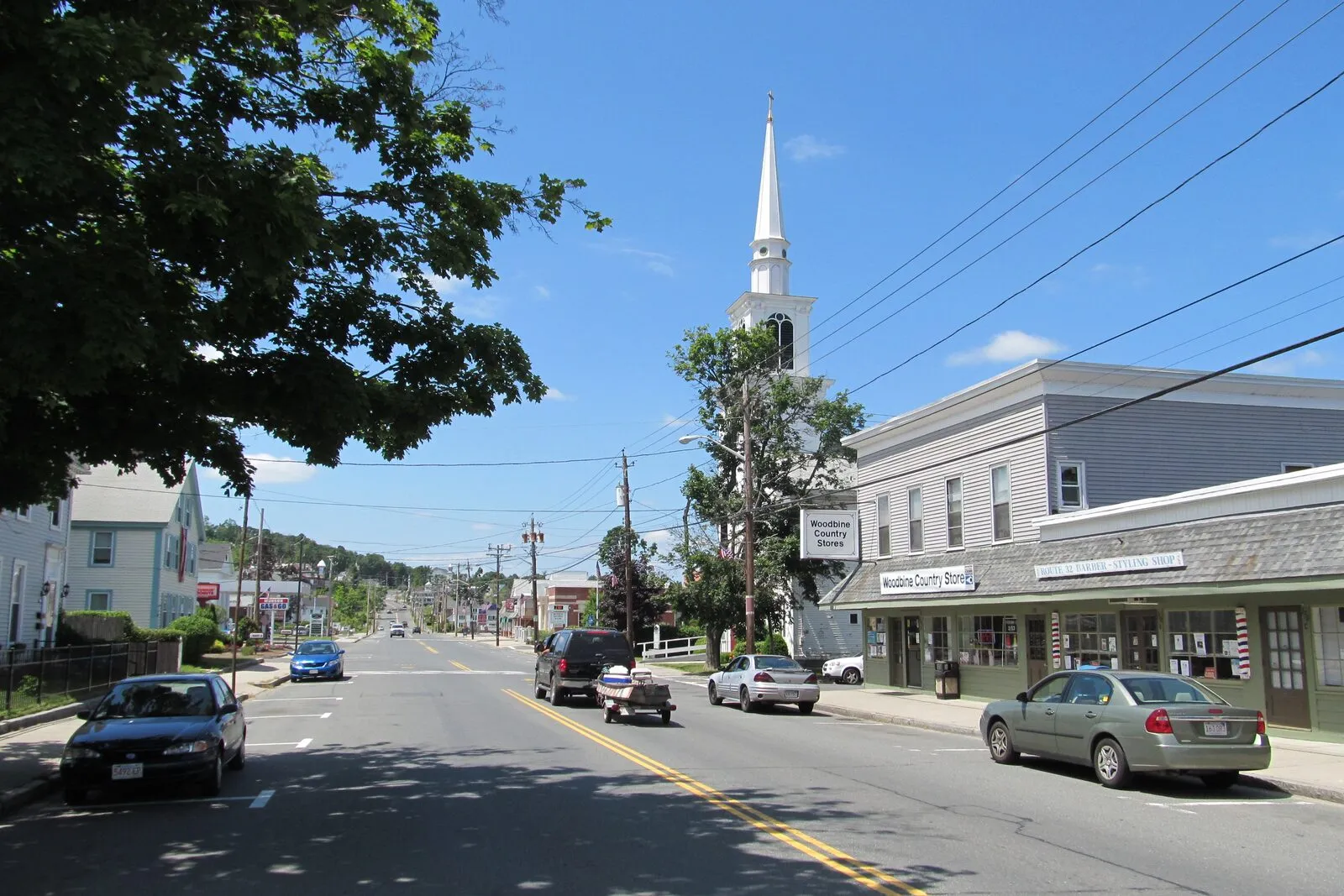 Looking south down Main Street (Route 32) in Monson on a clear summer afternoon: a two-lane village street with a tall white-clapboard Greek Revival church on the right, its slim white steeple rising above the surrounding rooflines, a low row of two-story commercial buildings beside it with painted storefront signs and a barber pole at the curb, a leafy maple arching in over the left side of the frame above clapboard houses set back behind a dark fence, parked sedans along the right curb, a few cars and a small utility trailer moving along the road, low forested hills closing the view at the far end of the street under a deep-blue sky scattered with light clouds.