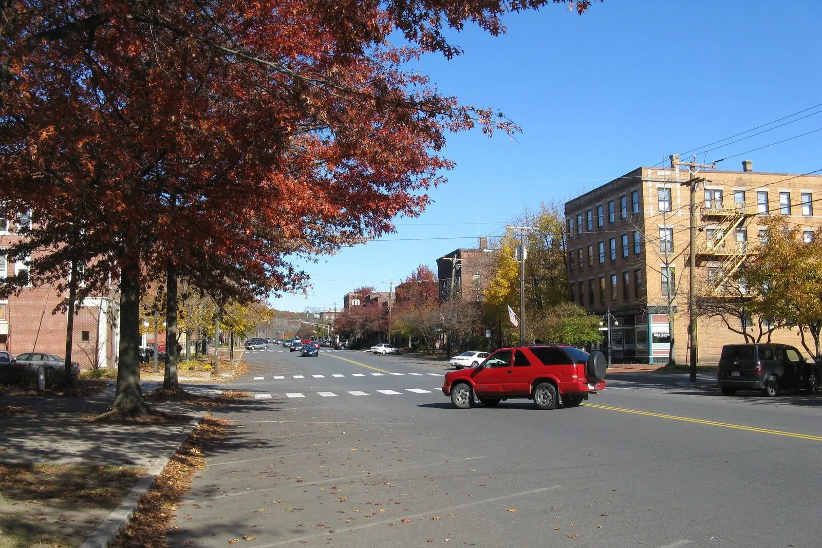 Avenue A in Turners Falls village in Montague on a clear autumn afternoon. A wide downtown street running through a row of late-19th-century brick commercial blocks on the right with mansard roofs and storefronts at street level, a row of mature trees in deep red and yellow fall color along the left sidewalk with leaves drifted onto the grass, a single red SUV crossing the street in the middle distance, low forested hills closing the view at the end of the street, and a clear blue sky overhead.