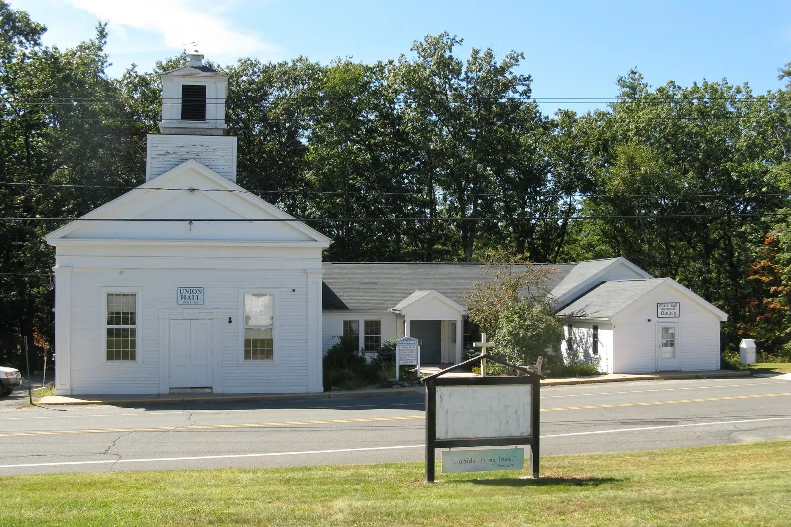 The Montgomery town center on a clear, sunny late-summer afternoon — a small white-clapboard Greek Revival meetinghouse with a square belfry and tall louvered cupola on the left, a sign reading 'Union Hall' over its front door, attached by a low connector to a one-story white town offices wing on the right with a small entry porch, dense green deciduous trees crowding in close behind the buildings, a two-lane road and a strip of mown lawn with a community bulletin board on tall posts running across the foreground, and a pale blue sky overhead.