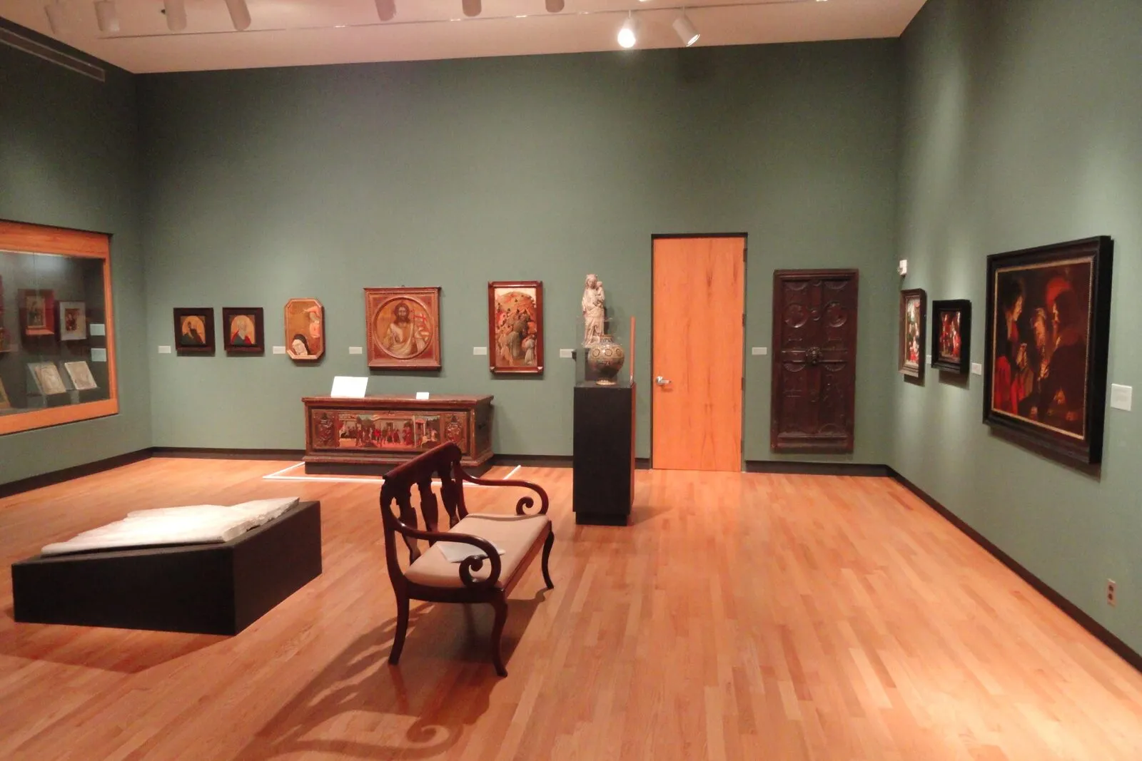 A medieval and Renaissance gallery at the Mount Holyoke College Art Museum: sage-green walls, light wood floor, track lighting overhead, four small early-Renaissance panel paintings on the left wall, a long polychrome wood altar chest in the center, a polychromed wood Madonna sculpture on a black pedestal beside it, a Caravaggesque oil painting on the right wall, a carved medieval wooden door panel mounted to the right of an interior door, a Federal-era settee on the floor, and a low display vitrine on the left.