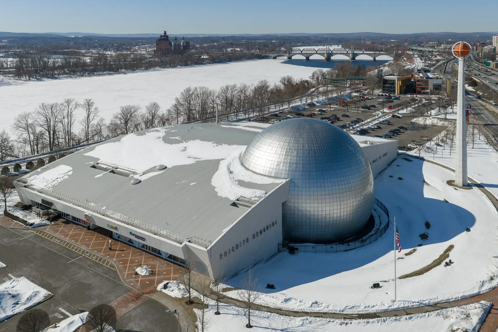 An aerial view of the Naismith Basketball Hall of Fame in winter. The chrome silver basketball dome at center, the flat-roofed rectangular museum wing extending to the left, a tower hoisting a giant red basketball on the right, flags below the tower, and a frozen Connecticut River with a highway bridge and the distant Springfield skyline behind.