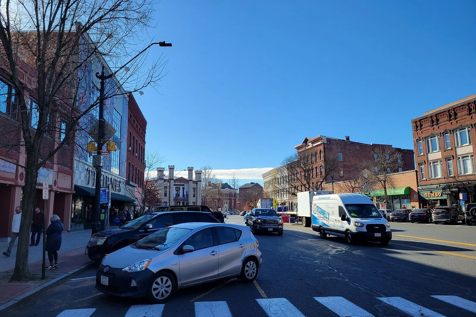 A view east along Northampton's Main Street on a bright November afternoon — the wide one-way street running across the frame with parked cars along both curbs, a continuous row of two- and three-story 19th-century brick and clapboard storefronts with painted-sign cornices on either side, mature street trees in late-fall foliage along the sidewalks, the Calvin Theatre marquee visible part-way down the block on the right, the white spire of the First Church rising above the rooflines in the distance, and a high blue sky overhead.