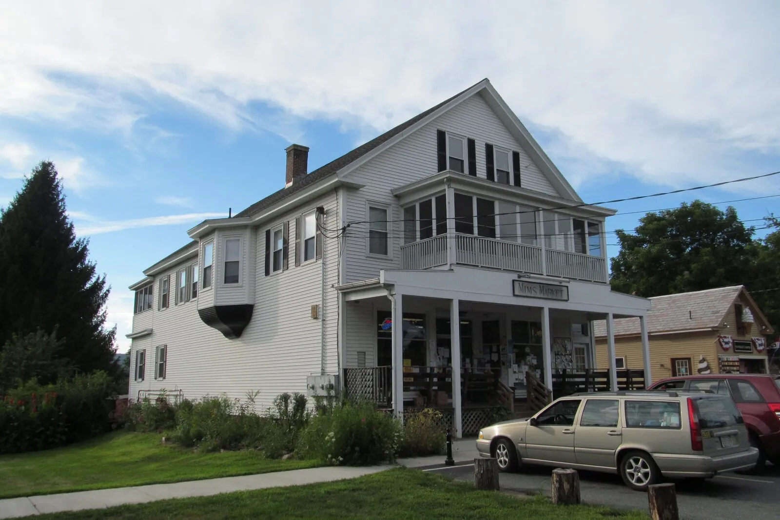 Mim's Market on Main Street in Northfield on a sunny late-summer afternoon. A tall white-clapboard 19th-century commercial building with a steep gabled roof, multiple gabled bays facing the street, a covered front porch with a hand-painted 'MIMS MARKET' sign over the entry, an upper-story sun porch with a railing, neatly planted gardens and shrubbery flanking the porch, a tall conifer hedge at left, a single tan station wagon parked at the curb in front, an adjacent Cape-style house barely visible to the right, mature green trees beyond, and a pale blue sky scattered with thin clouds.