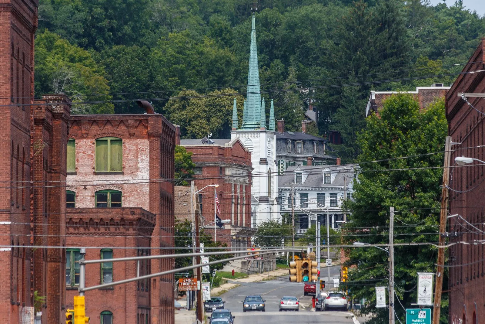Looking north along South Main Street in downtown Orange on a clear summer afternoon — a tall four-story red-brick commercial block at the left foreground with rows of arched windows and a stepped cornice, the white-painted Central Congregational Church with its slender green-trimmed Gothic spire rising in the middle distance, white-clapboard 19th-century commercial and residential buildings down the right side of the street, overhead utility wires crossing the view, traffic lights and a yellow Route 122 marker over an empty roadway, a few cars parked and approaching, and forested low hills closing the view at the end of the street under a hazy blue sky.