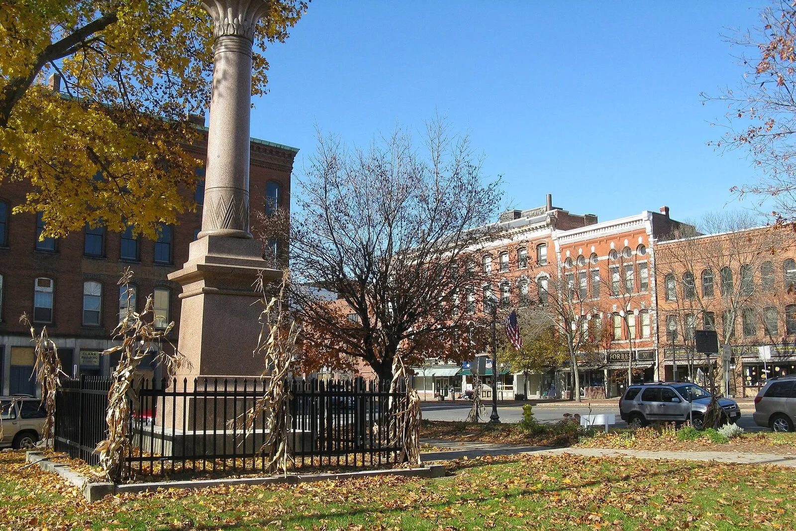 Court Square in downtown Greenfield on a clear November afternoon. A small triangular town square paved in granite-curbed concrete with a low fountain at the center, the brownstone-and-granite Franklin County Courthouse rising on the far side with its tall central tower and slate roof, a row of 19th-century three-story brick commercial buildings (including the block holding the People's Pint a short walk down Federal Street) facing the square on the right, bare-branched mature trees ringing the open ground, and a clear pale blue sky overhead.