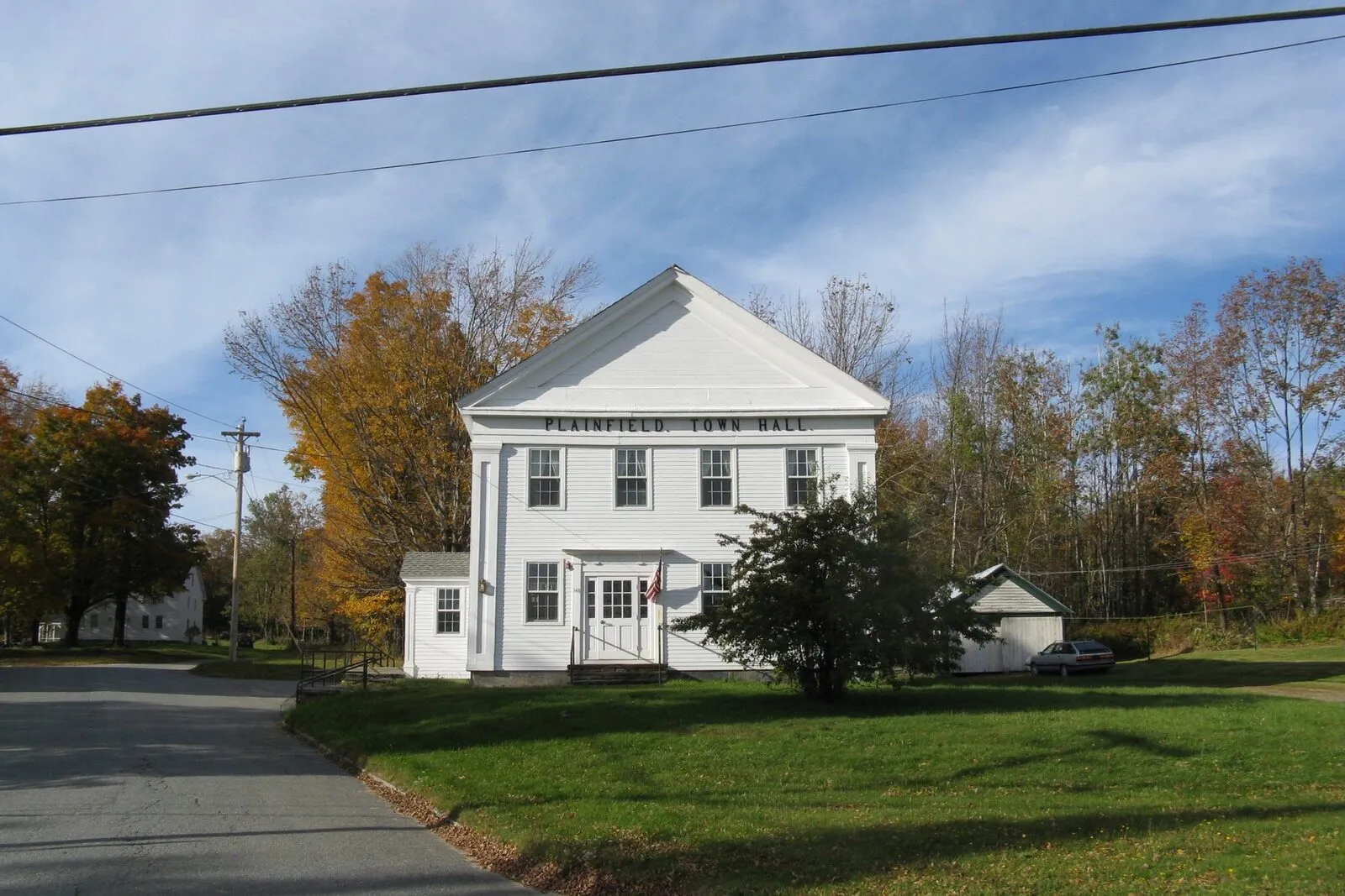 Plainfield Town Hall on a clear fall afternoon: a two-story white-clapboard Greek Revival meetinghouse with a steep gabled roof forming a full pediment across the front, the words PLAINFIELD TOWN HALL painted in dark lettering across the pediment frieze, a single central entry door reached by a short flight of stairs with a black handrail, two narrow rectangular windows flanking the door, four tall sash windows on the upper story, a small low side wing to the right with a metal standing-seam roof, a dark green shrub beside the steps, mature deciduous trees in mixed orange-yellow-and-green fall foliage framing the building, a parked dark sedan barely visible past the right edge, a quiet two-lane road curving in the foreground, and a deep blue sky with thin scattered cirrus clouds overhead.