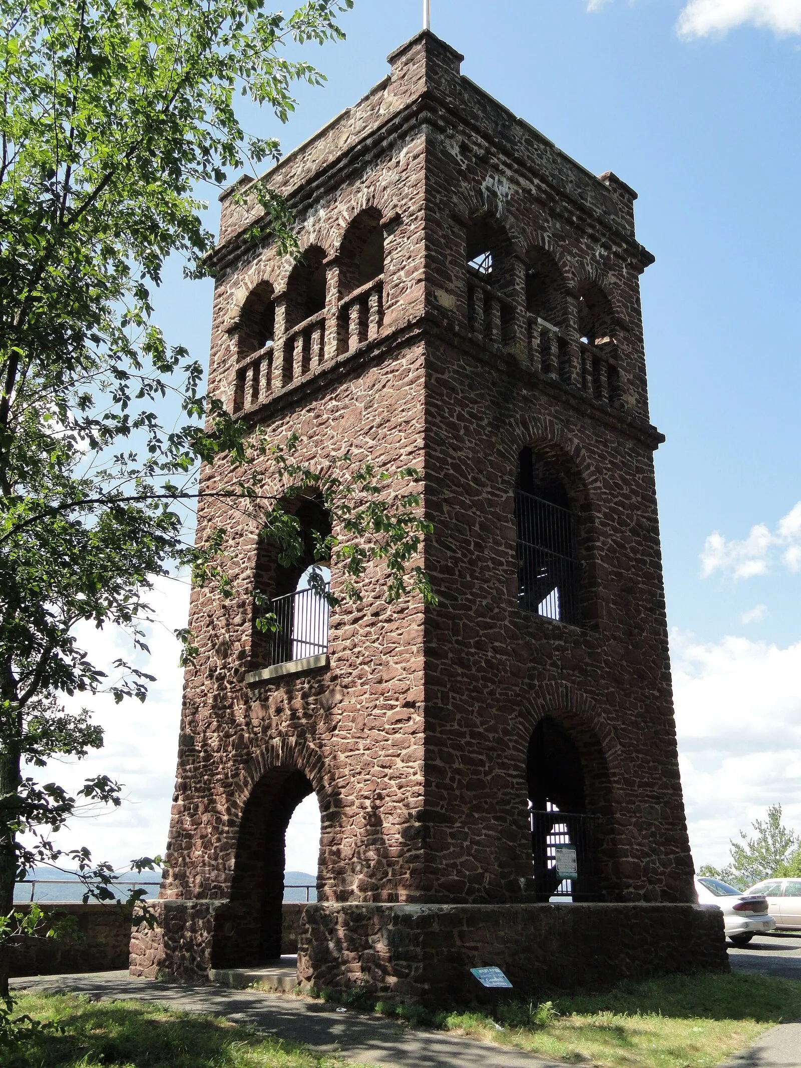 Poet's Seat Tower on a sunny summer afternoon, a square red-sandstone observation tower with a crenellated battlemented top, three stories of arched window openings, and an arched entry at ground level, framed by green deciduous foliage on the left, with a sliver of the valley visible behind on the right under a clear blue sky with light cumulus.