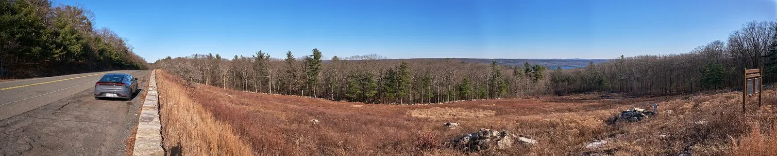 Wide panorama from the Pelham Overlook pull-off on Route 202 in Pelham on a clear winter afternoon — a parked dark-gray hatchback at the left edge of the asphalt shoulder, a low stone retaining wall along the road, a broad expanse of brushy meadow and bare deciduous forest below, a band of low forested hills rolling east toward the distant Quabbin Reservoir basin, the reservoir itself just visible as a thin pale line on the horizon at right, an interpretive sign on a wooden post at the far right, and a clear blue sky overhead.