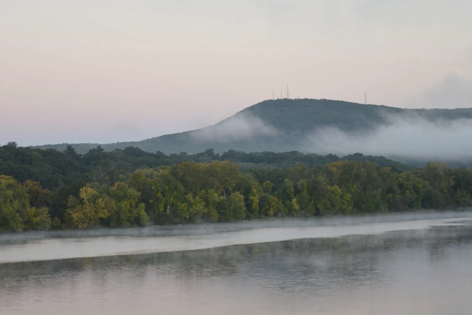 Mount Tom rising above a bank of fog, with the Connecticut River in the foreground — ribbons of mist lifting off the water, green trees along the far bank, and communications towers faintly visible on the summit.