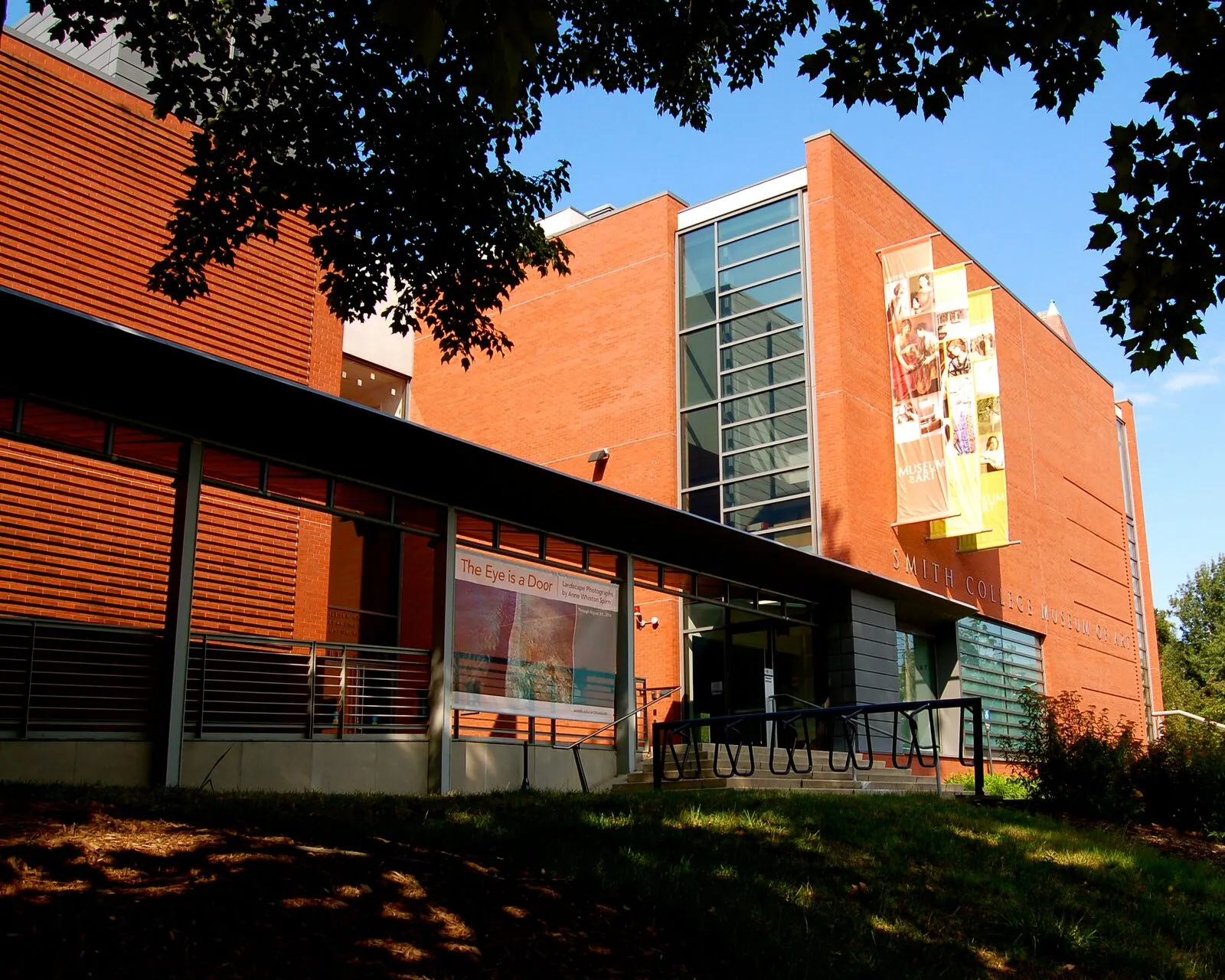 The Smith College Museum of Art entrance on Elm Street on a bright morning. A modernist red-brick building with a low horizontal-louvered wing on the left, a glass-walled stairwell in the center, and a taller brick pavilion on the right carrying a large vertical banner of an orange-and-yellow painted figure; the 'SMITH COLLEGE MUSEUM OF ART' wordmark runs above the glass doors, a small exhibition poster for 'The Eye Is a Door' hangs on the wing wall, and leafy tree branches frame the edges under a clear sky.