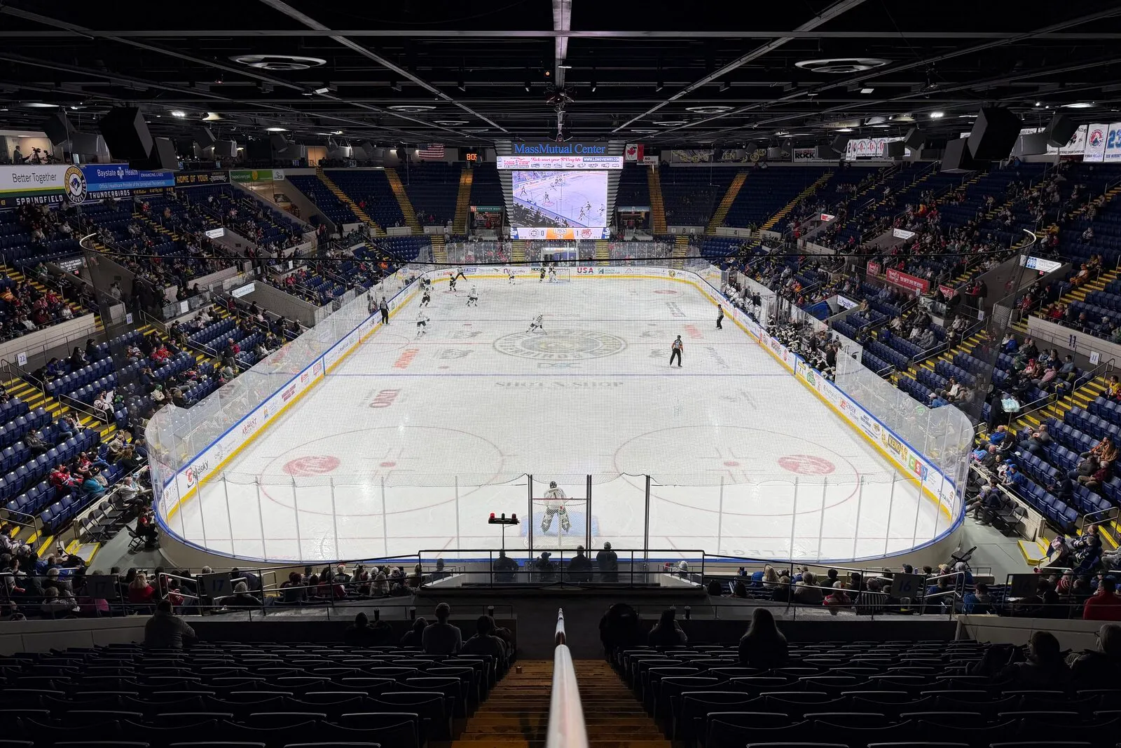 The interior bowl of the MassMutual Center in Springfield seen from an upper seating row before a Thunderbirds game on a fall evening. Bright white ice with red and blue lines, a center-ice logo, two faceoff circles in the foreground, players in dark and white sweaters skating in pre-game warmups, a four-sided overhead video scoreboard hanging above center ice, a curved upper deck with rows of dark-blue seats wrapping the rink, advertising boards along the dasher and on the rink-end walls, exposed steel ceiling trusses with stage lighting, and the empty backs of seats in the foreground row.