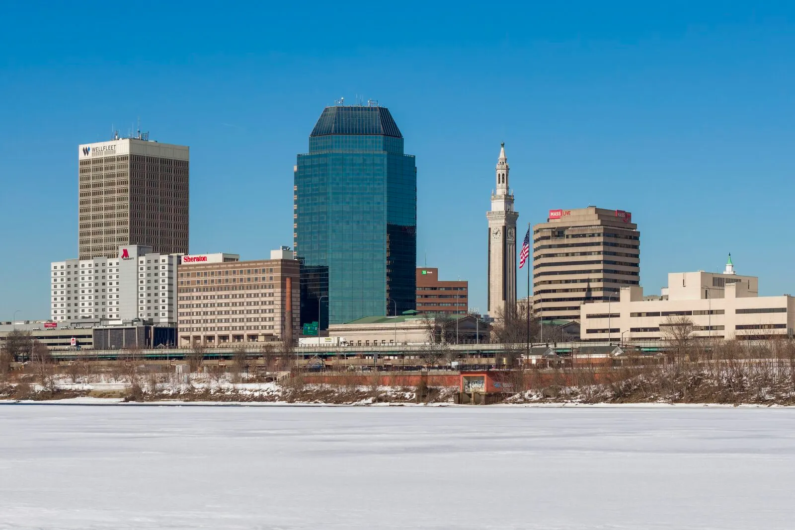 The Springfield skyline seen from across a frozen Connecticut River under a clear blue sky: the green-glass Monarch Place tower at center, the white Campanile bell tower of the Municipal Group rising beside it with a U.S. flag flying, a Sheraton and the 1550 Main building flanking the group, and low downtown blocks stretching along a snowy riverbank.
